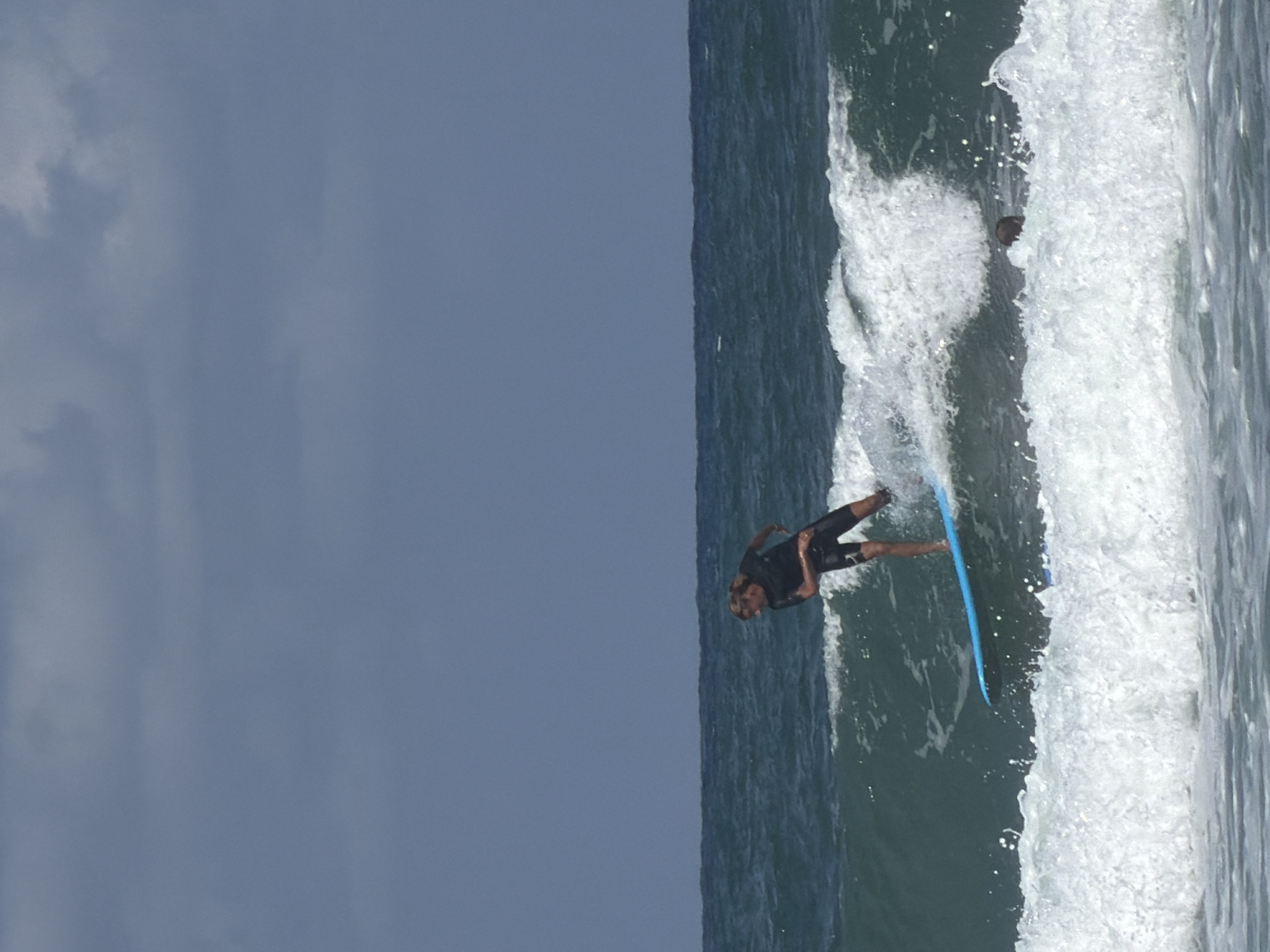 A surfer rides a wave off the coast of Tel-Aviv, Israel. The surfer, wearing a wetsuit, is riding a blue surfboard. The ocean waves are breaking. The sky is overcast. Another individual's head is visible behind the surfer.