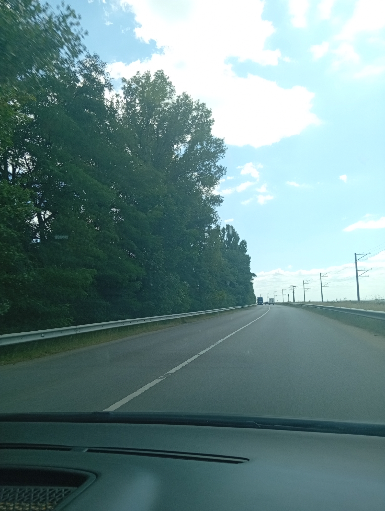 A car's perspective shows a clear blue sky with scattered clouds above a highway lined with tall green trees on the left. A metal guardrail runs along the side of the road. In the distance, power lines with tall poles are visible. Several vehicles, including trucks and cars, are driving on the highway. The scene appears to be in a rural or suburban area outside of a city.