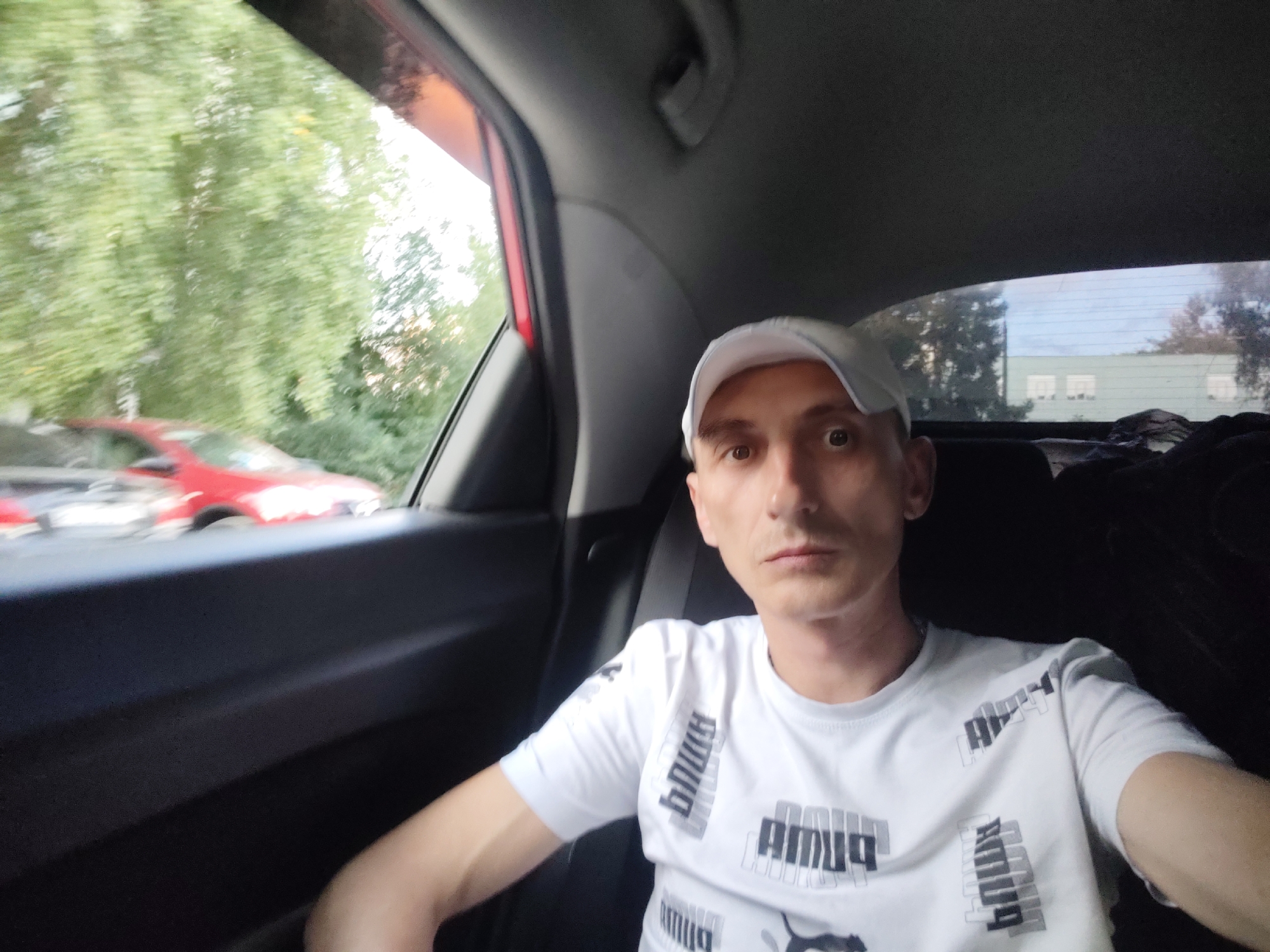 A man wearing a white baseball cap and a white t-shirt with a repeated "Puma" logo print is sitting in the back seat of a car. The man is looking directly at the camera with a neutral expression. The interior of the car is visible, including the dark upholstery of the seats and the car's ceiling. Through the car window, lush green trees and a blurry red car are visible, suggesting the car is in motion or parked near a vegetated area. The sky appears bright, hinting at daylight. In the distance, through the rear window, a building with multiple windows can be seen. The scene appears to be set in Bor, Russia. There is no other discernible activity or event taking place. The visible text on the man's t-shirt is "Puma".