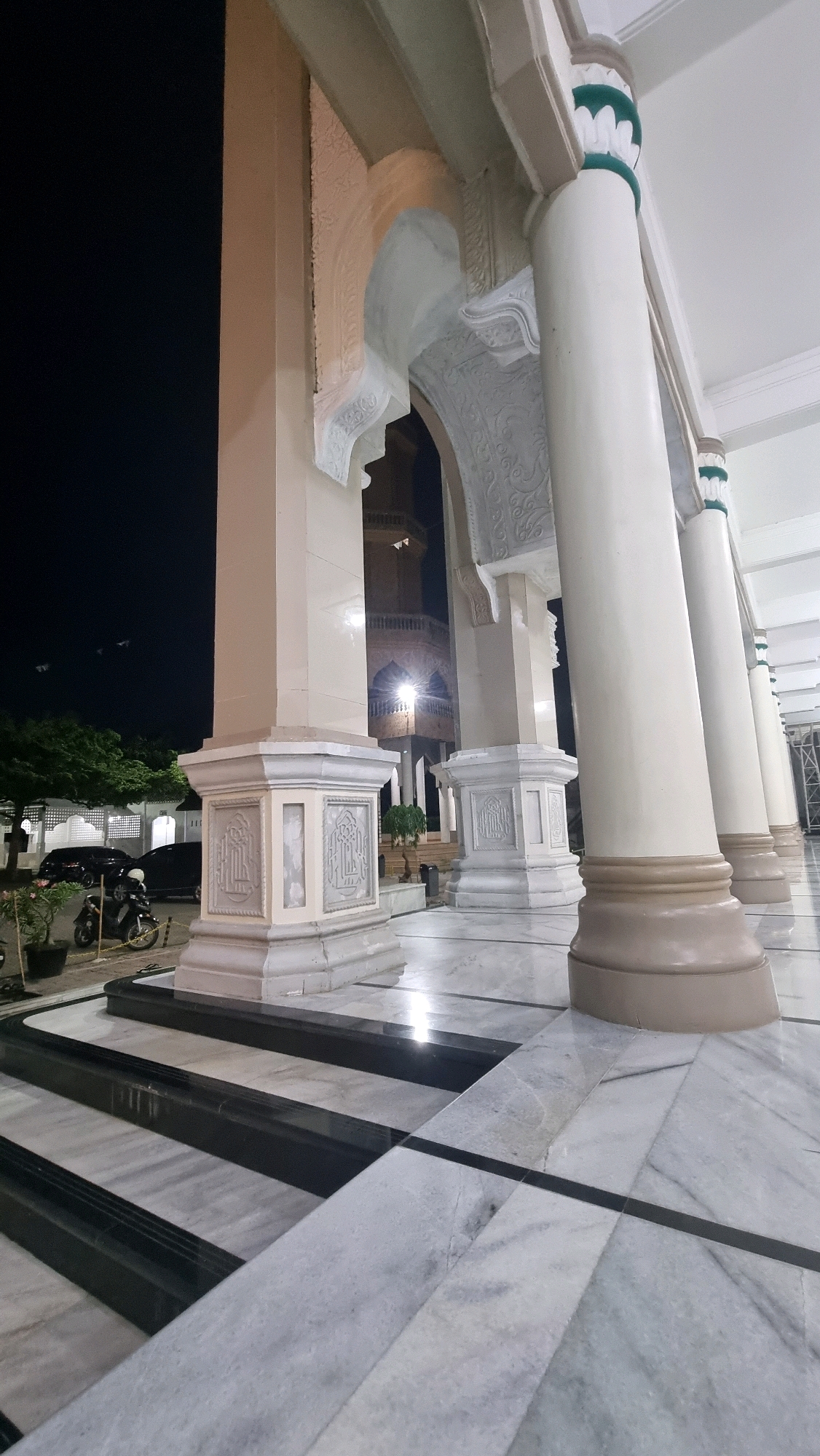 A low-angle, night-time, exterior shot of a mosque in Lhokseumawe, Indonesia, showcases a portion of its facade. The foreground is dominated by a marble floor with black accent lines, leading up to a series of large, cream-colored pillars. These pillars are ornate, with detailed carvings at their bases. Behind the pillars, an arched entryway reveals a dimly lit tower, characteristic of a mosque's minaret. Trees and parked vehicles, including a motorcycle and two cars, are visible in the background, illuminated by artificial lights against the dark night sky.