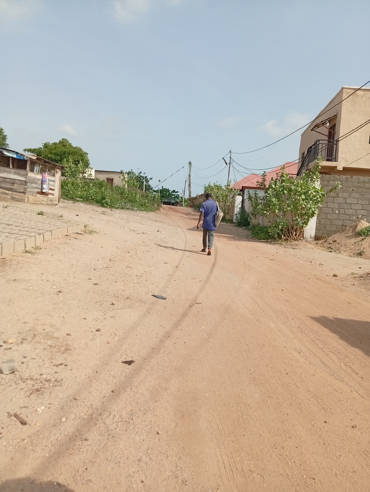 The image depicts a dusty, unpaved road in Adankolo, Nigeria. A man is walking away from the camera, down the center of the road. He is wearing a blue shirt and dark pants, and appears to be carrying a bag. The road is lined with buildings of varying construction styles and materials. The buildings consist of a structure of wood, a house with a red roof, and a structure built of brick. Power lines run overhead. Vegetation is present along the roadside. The sky is light blue with scattered clouds.