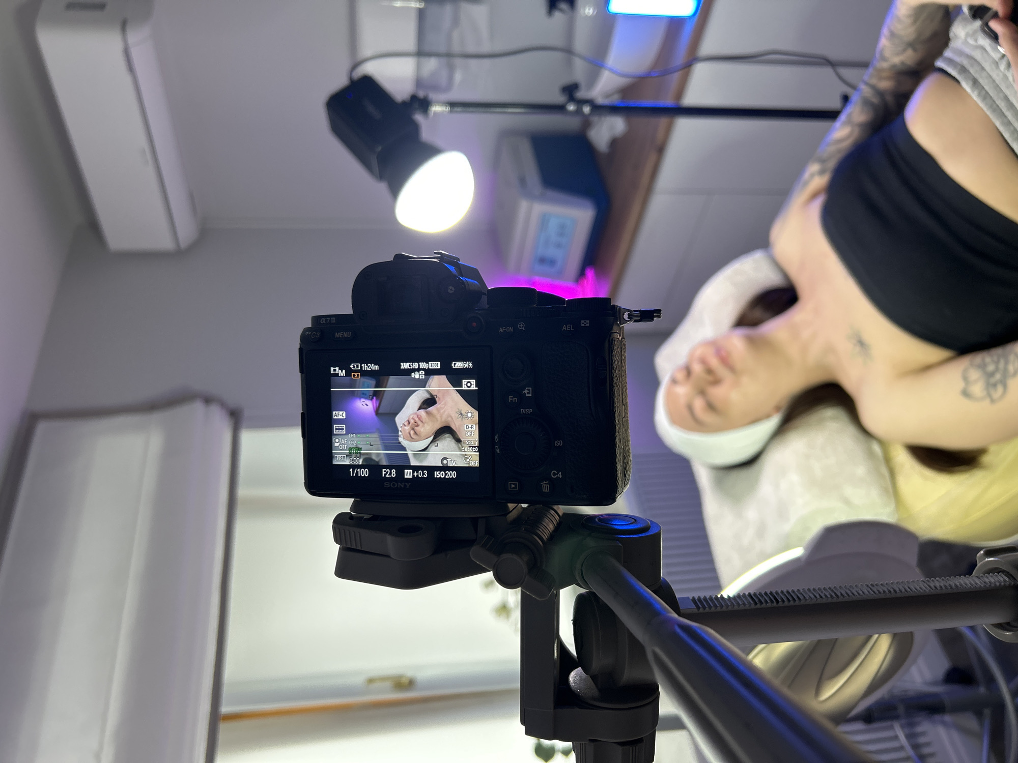 A close-up, indoor shot shows a Sony α7 III camera mounted on a tripod, focused on a person lying on a padded surface. The camera screen displays the image it is capturing, showing the person's face and upper body. The camera settings visible on the screen are: shutter speed 1/100, aperture F2.8, exposure compensation +0.3, and ISO 200. The camera mode is set to Manual (M) and autofocus is continuous (AF-C). A video light on a stand provides illumination for the shot. The scene appears to be a photography or videography setup for capturing content of a person, possibly related to beauty or wellness. The location is not discernible from the image and therefore a specific location in Votkinsk, Russia cannot be determined.