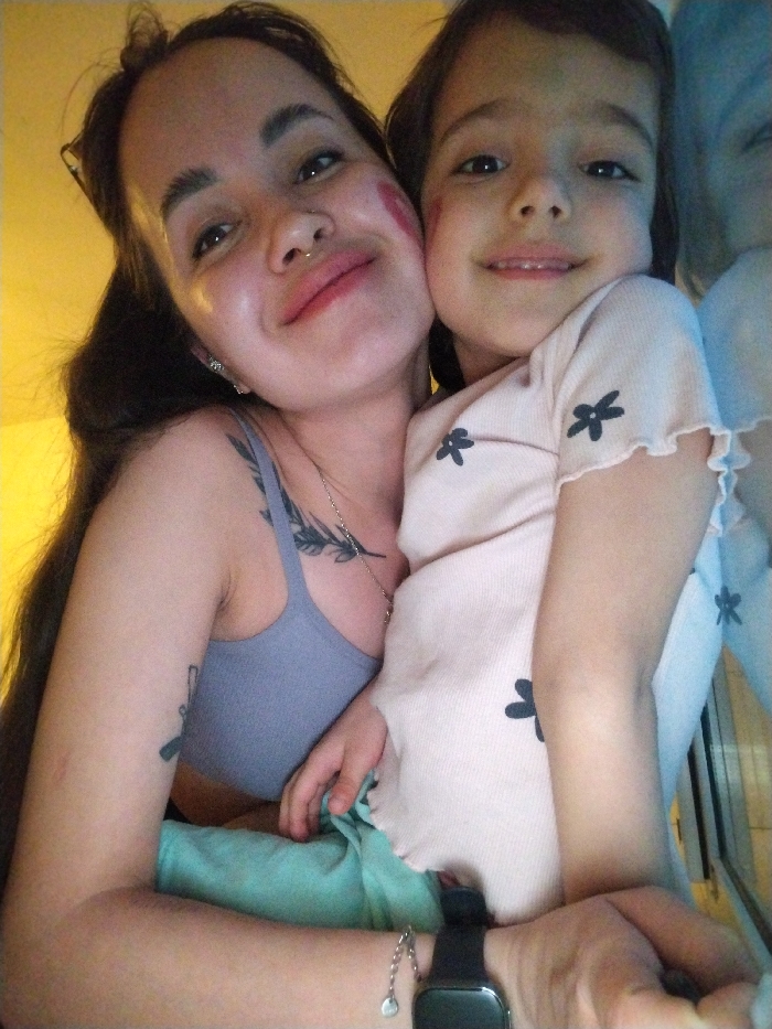 The image depicts two individuals, an adult female and a young child, embracing and smiling in Noviy, Russia. The adult female has long dark hair, light skin, and is wearing a light blue bra-top. She exhibits a gold-toned septum piercing, an ear piercing, glasses pushed up on her head, and two tattoos: a leafy branch design on her upper left chest and a rifle-like silhouette on her left bicep. A red kiss mark is visible on her left cheek. She wears a black smartwatch and a silver chain bracelet with a circular charm on her left wrist. The child, appearing to be a young girl with brown hair and light skin, is wearing a light pink ribbed top with dark flower patterns and light green pants. A red kiss mark is present on her right cheek. The adult female's left arm is wrapped around the child, who is positioned to the adult's left side. Both subjects are looking directly at the camera. The background is indistinct; a warm yellow tone is visible on the left, and a bluish area with a faint reflection and a striped pattern is present on the right.