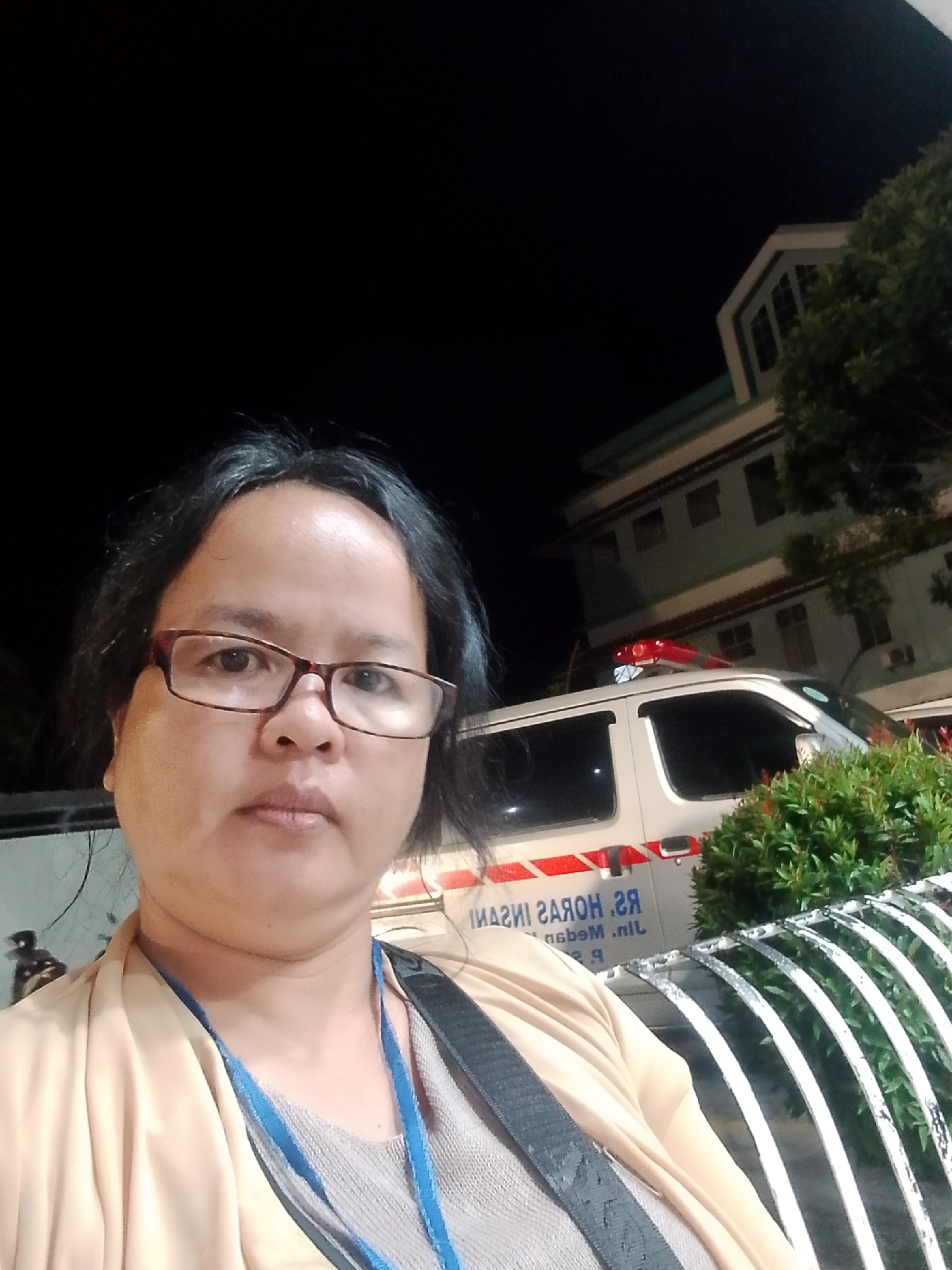 A medium shot selfie depicts a woman wearing eyeglasses and a beige cardigan over a gray shirt, with a blue lanyard visible. She is positioned in front of a building with an ambulance parked to her right. The building appears to be a hospital or medical facility, indicated by the presence of the ambulance. The ambulance has red and white markings, and a red emergency light bar is mounted on its roof. In the background, a portion of a tree is visible, and the scene is illuminated by artificial light, suggesting it is nighttime. Text on the ambulance is partially visible and appears to be in Indonesian. The overall setting is outdoors, possibly a parking lot or entrance area of a medical facility.