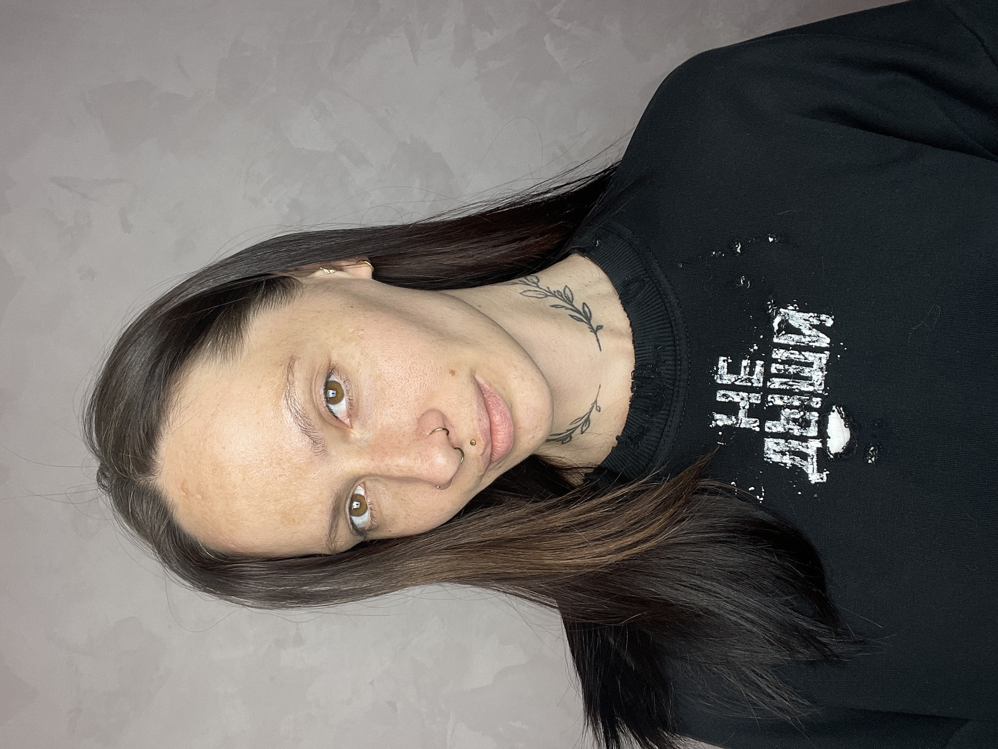 A portrait-style image, rotated vertically, features an adult female with long, dark, straight hair flowing over her right shoulder and across the black garment she is wearing. Her head is tilted, and she is looking directly at the viewer with light brown eyes. Her complexion is fair with visible freckles across her nose and cheeks.
She has several facial piercings: a gold-colored septum ring in her nose and a small gold stud above her upper lip, just off-center. A minimalist tattoo of a leafy branch design is visible on the right side of her neck, trailing from behind her ear towards her collarbone. Another small gold-colored earring is visible in her right earlobe.
She is wearing a black long-sleeved top or sweatshirt, which has a distressed, white graphic print on the front. The text in the graphic is partially obscured and difficult to decipher but appears to be in a stylized, somewhat grunge font. Her hair falls partly over the left side of the graphic.
The background is a plain, light grey wall with a subtle, irregular texture, suggesting an indoor setting. The lighting is soft and even, highlighting her features without harsh shadows. The overall mood of the image is calm and direct. The location context for this media file is Izhevsk, Russia.