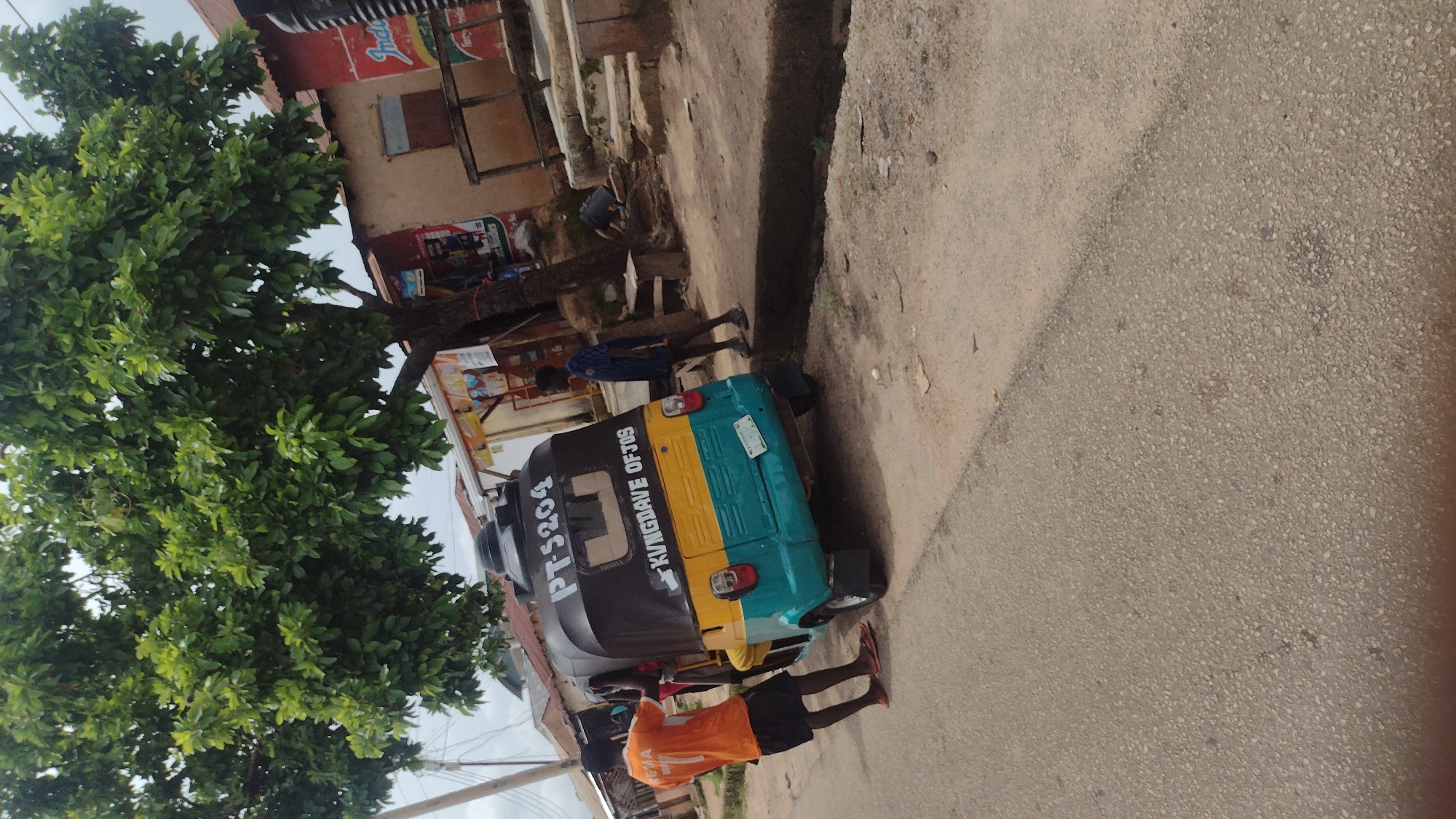 The image depicts a street scene in Jos, Nigeria, under an overcast sky. In the foreground, a three-wheeled vehicle, commonly known as a Keke Napep, is parked on the right side of a paved road. The Keke Napep has a black canopy with white lettering that reads "PT5204" and below that, "KINGDAVE OF JOS". The lower portion of the vehicle is painted in yellow and turquoise. Two young men are interacting with the Keke Napep. One man, wearing an orange t-shirt with the number "77" and black shorts, stands to the left of the vehicle, facing it and appearing to be engaged in conversation or some activity with the occupants. Another young man, wearing a blue and black patterned t-shirt and dark shorts, stands further back on the right, observing the scene.
The environment suggests a commercial or residential area within Jos. A large tree with dense green foliage dominates the upper left portion of the image. Behind the Keke Napep and to the right, there are several small buildings with corrugated iron roofs, typical of structures found in many Nigerian cities. Some of these buildings appear to be shops, with signs and posters visible on their facades. A wooden structure, possibly a stall or part of a shop, is situated in front of one of the buildings. The road itself is asphalted, with a visible drainage channel running alongside it on the right. The overall impression is of a typical street in a bustling, developing urban environment.