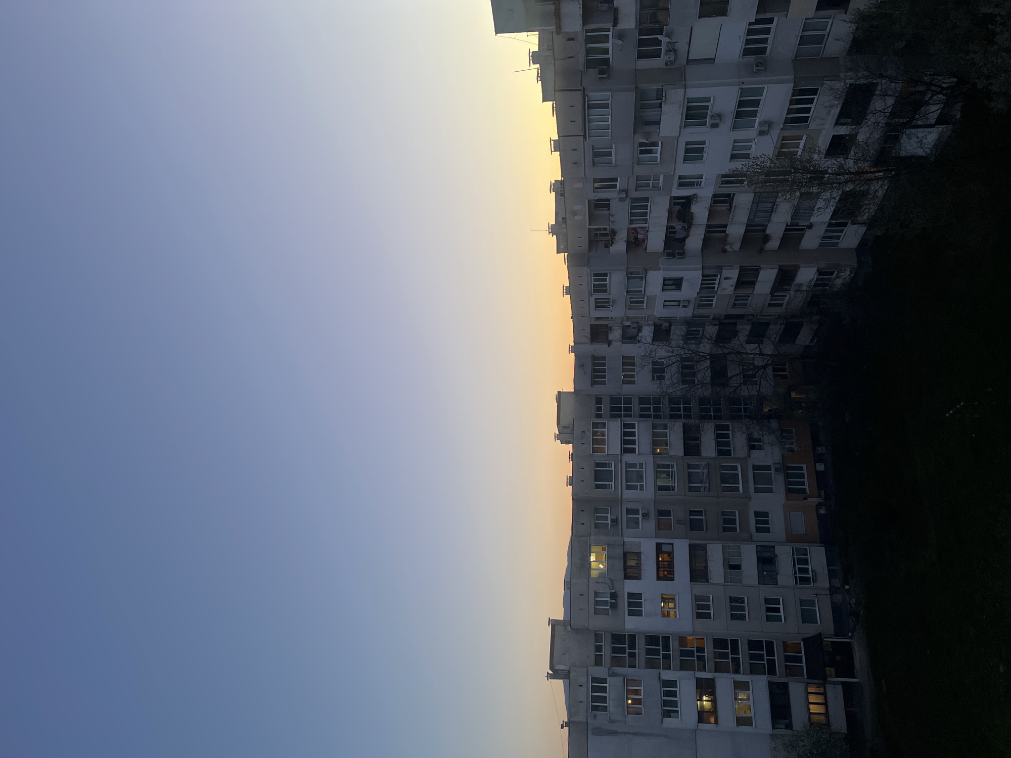 The image shows a residential building in Sofia, Bulgaria, viewed from a low angle. The building is concrete, multi-storied, and features many windows, some with lights on. The sky above transitions from blue to orange. The lighting suggests it is either dawn or dusk. Trees are visible on the right edge of the frame. No people are visible.