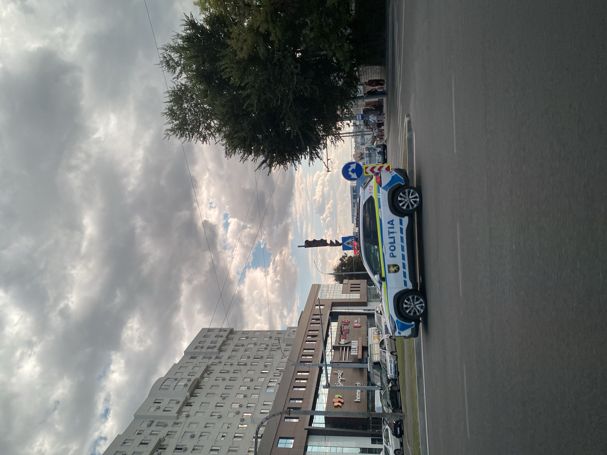 The image depicts a street scene in Chișinău, Moldova, under a cloudy sky. A white police car with blue and yellow markings, displaying the word "POLITIA," is prominent in the foreground, positioned on the right side of the road, facing left. The police car has a symbol on its side that appears to be a coat of arms. Behind the police car, several other vehicles, including cars and a van, are visible on the road.
In the background, a large, multi-story white building with many windows dominates the left side of the frame. Below this building is a commercial establishment with signage that includes "Simpals" and "RE/MAX." To the right of the commercial building, a traffic light with red and yellow signals is visible. Further to the right, a large tree with dense foliage stands near the edge of the frame.
Several individuals are seen on the sidewalk near the tree and the road, with some appearing to be waiting. A pedestrian crossing is marked on the road in front of the people. A blue circular traffic sign with a white arrow indicating a turn is also visible. The overall environment suggests an urban setting, likely during daylight hours, with the weather characterized by scattered clouds.