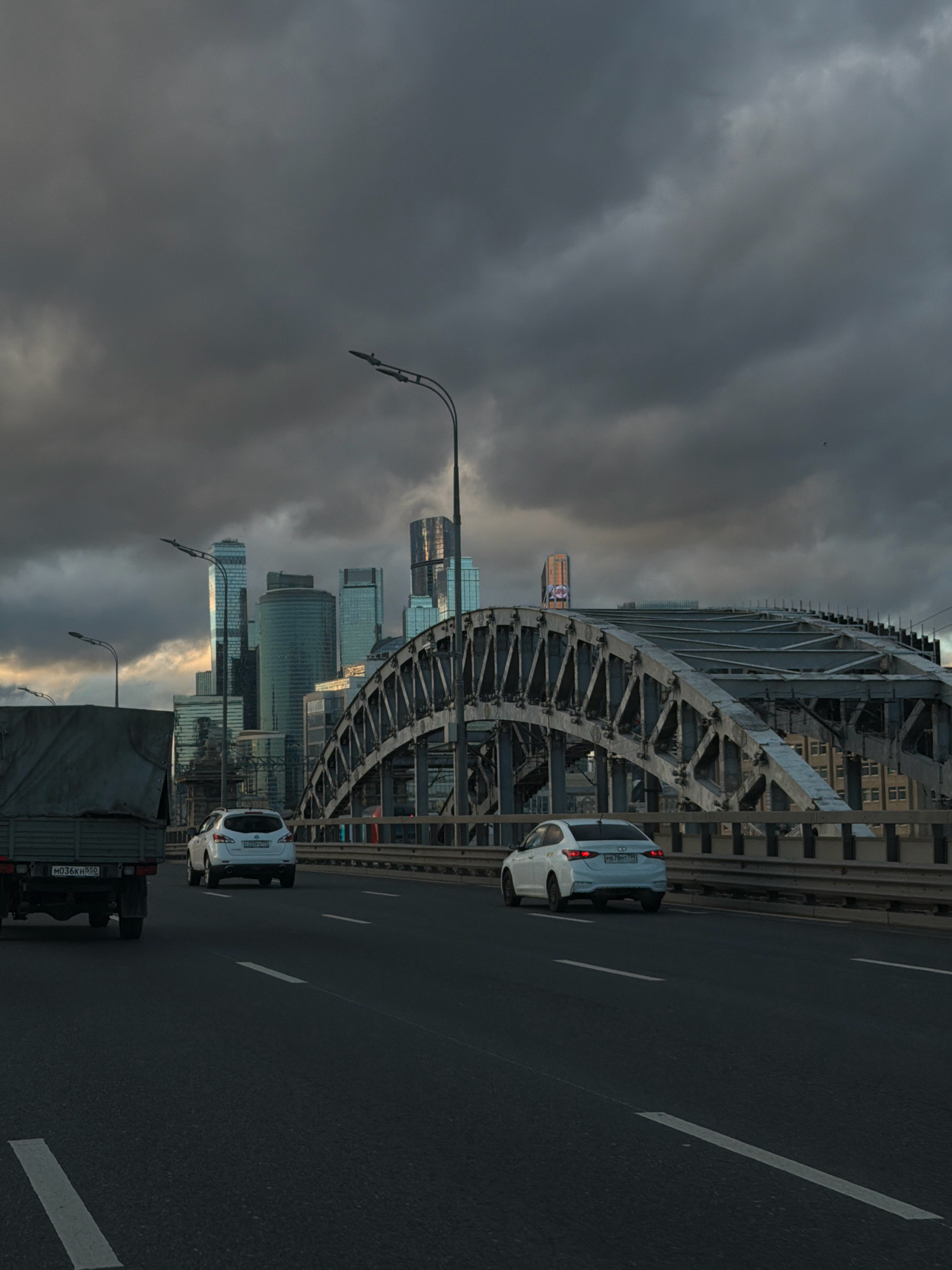 A low-angle, wide shot captures a dramatic view of a highway bridge in Moscow, Russia, under a heavily overcast sky. The foreground shows a dark asphalt road with white lane markings. Two white cars are driving on the bridge, one in the left lane and another in the right lane, further ahead. The car in the left lane is closer to the viewer, a white SUV. The car in the right lane is a white sedan. To the left of the road, a large, gray truck with a covered bed is visible. The middle ground is dominated by the arched structure of the bridge, made of steel beams. In the background, the Moscow skyline is visible, featuring numerous modern skyscrapers with glass facades, reflecting the dim light. The sky is filled with dark, ominous clouds, creating a somber and dramatic atmosphere.