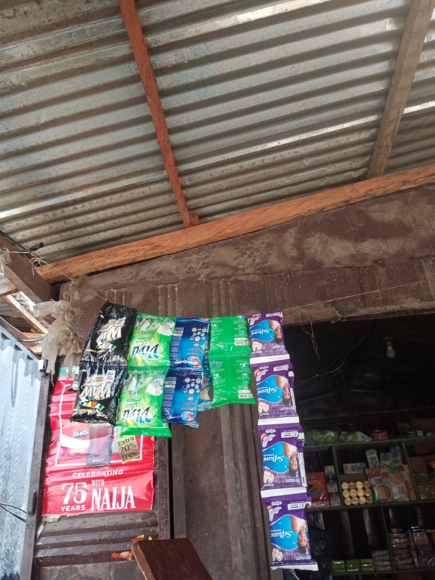 The image shows an outdoor stall in Jalingo, Nigeria, selling various consumer goods. The stall appears to be covered by a corrugated metal roof supported by wooden beams. Hanging from a wire, a variety of packaged items are displayed, including detergent packets and what appear to be sanitary pads. A large red banner is visible in the foreground, featuring text that reads "CELEBRATING WITH 75 YEARS NAIJA". In the background, shelves are stocked with additional products, including boxes of Kellogg's Corn Flakes and other packaged foods. The overall impression is of a small, informal retail space.