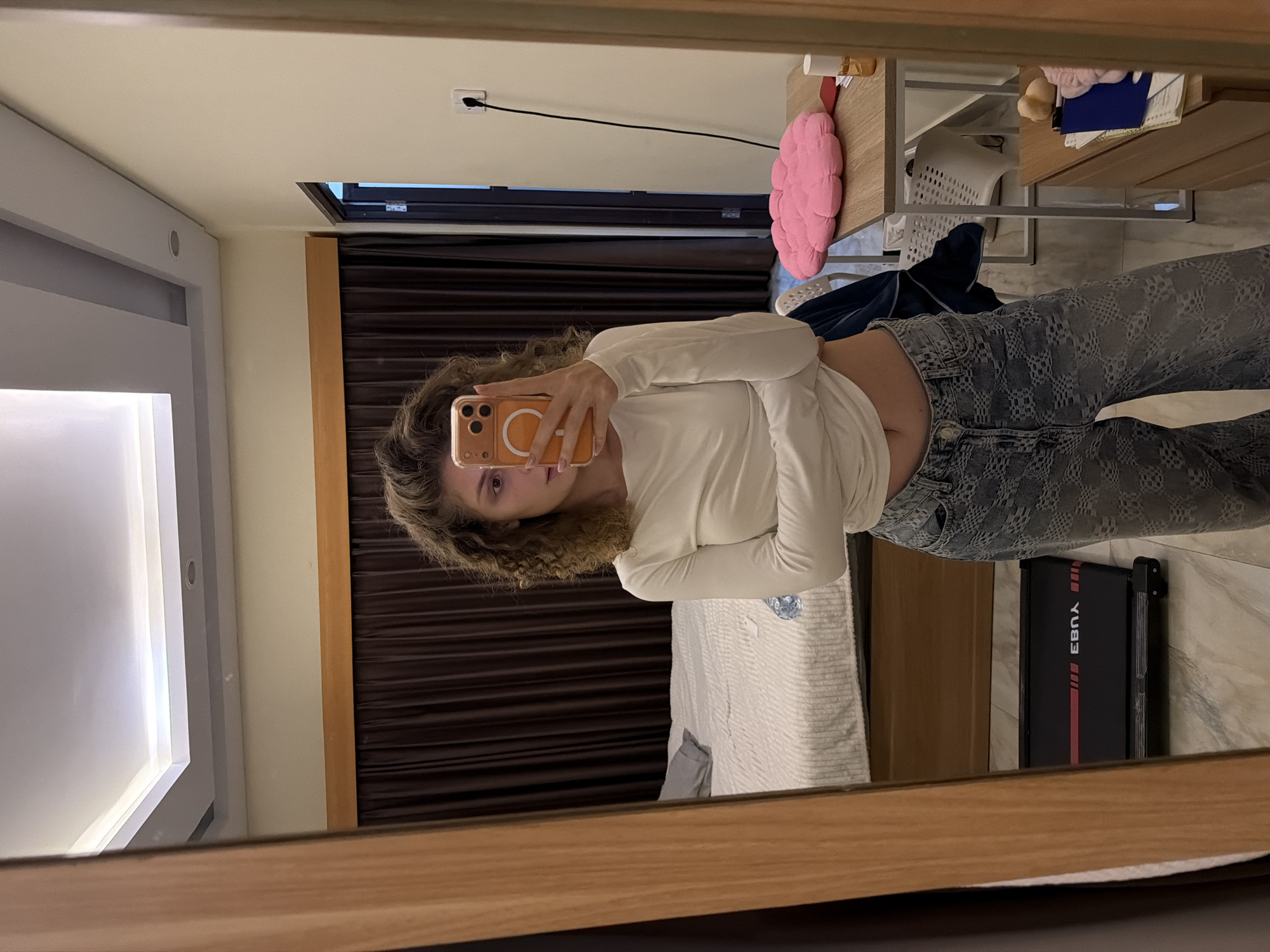 A woman with curly hair is taking a selfie in a mirror. She is wearing a long-sleeved white shirt and gray patterned jeans. The mirror reflects a room with brown curtains, a bed with white linens, a gray treadmill, and a desk. There is a pink pillow on the desk. The room is located in North Kuta, Indonesia.