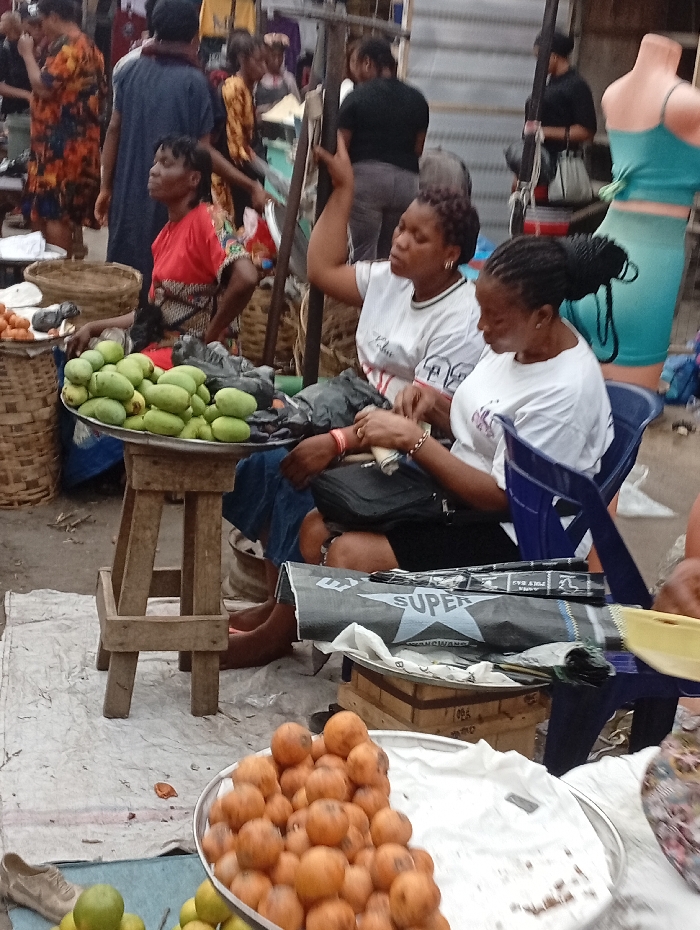 A bustling market scene shows two women seated, selling produce. One woman, wearing a red top, sits beside a tray piled high with green mangoes. Adjacent, two women in white t-shirts sit with a second tray displaying orange fruits. A backdrop of people, clothing, and wares fills the frame, creating a vibrant, busy atmosphere. Baskets and a mannequin add to the commercial tableau.