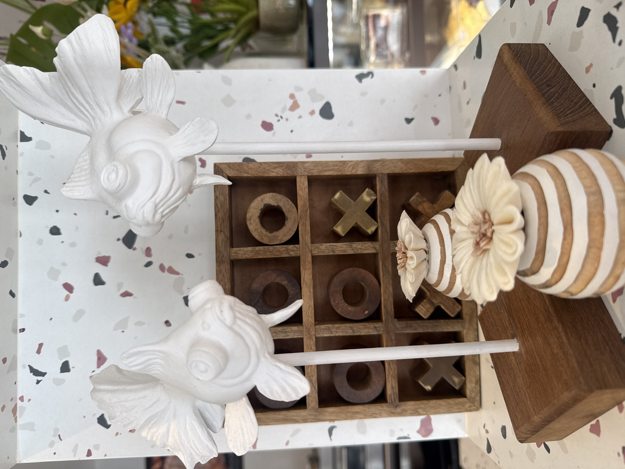 A white ceramic fish sculpture is mounted on a white stick, which is inserted into a dark wood base. The sculpture is in the foreground, slightly to the left of the image center. Behind it, a wooden tic-tac-toe board is visible. Some wooden 'O' pieces and gold-colored 'X' pieces are placed in the squares. To the right and left of the tic-tac-toe board are two striped decorative vases with flower-like tops. In the background, a wall with a terrazzo pattern is visible, and to the far right, a blurred arrangement of greenery and flowers can be seen, along with a glimpse of display shelves, suggesting this scene is likely in a shop or cafe in Ko Samui District, Thailand.