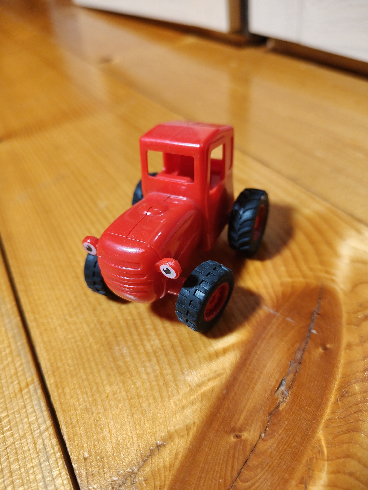 A bright red toy tractor with black wheels and painted-on eyes sits on a wooden surface. The tractor is positioned at an angle, with its front facing towards the viewer's left. The wooden surface appears to be a table or floor made of planks with visible grain. The background is blurred, showing a light-colored wall or furniture. The scene is well-lit, casting a shadow of the tractor to its right.