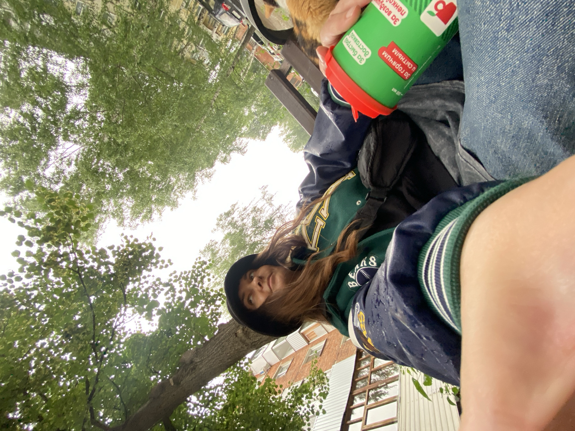 This is a vertical, wide-angle, eye-level selfie taken outdoors, likely in Votkinsk, Russia, during the daytime. The sky is overcast, suggesting a cloudy or rainy day. The scene is dominated by lush green trees with visible branches and leaves, creating a canopy overhead. In the foreground, a person is captured from a low angle, wearing a dark-colored baseball cap and a green and navy blue jacket with yellow accents. The jacket appears wet, possibly from rain. The person's long, brown hair is visible, and they are looking towards the camera with a neutral expression.
To the right of the person, a portion of another person's blue denim jeans is visible. The person taking the selfie is holding a bright green cup with a red lid. The cup has text on it. Some visible text on the cup, in Cyrillic script, includes "За быстрый" and "За воздушным". There is also a small red apple icon. In the background, there are hints of a building with windows and wooden structures, suggesting an urban or suburban environment. A small section of what appears to be an orange cat's fur is visible near the top of the frame, held by the person taking the photo, suggesting interaction with a pet. The overall impression is a casual, candid moment captured outdoors.