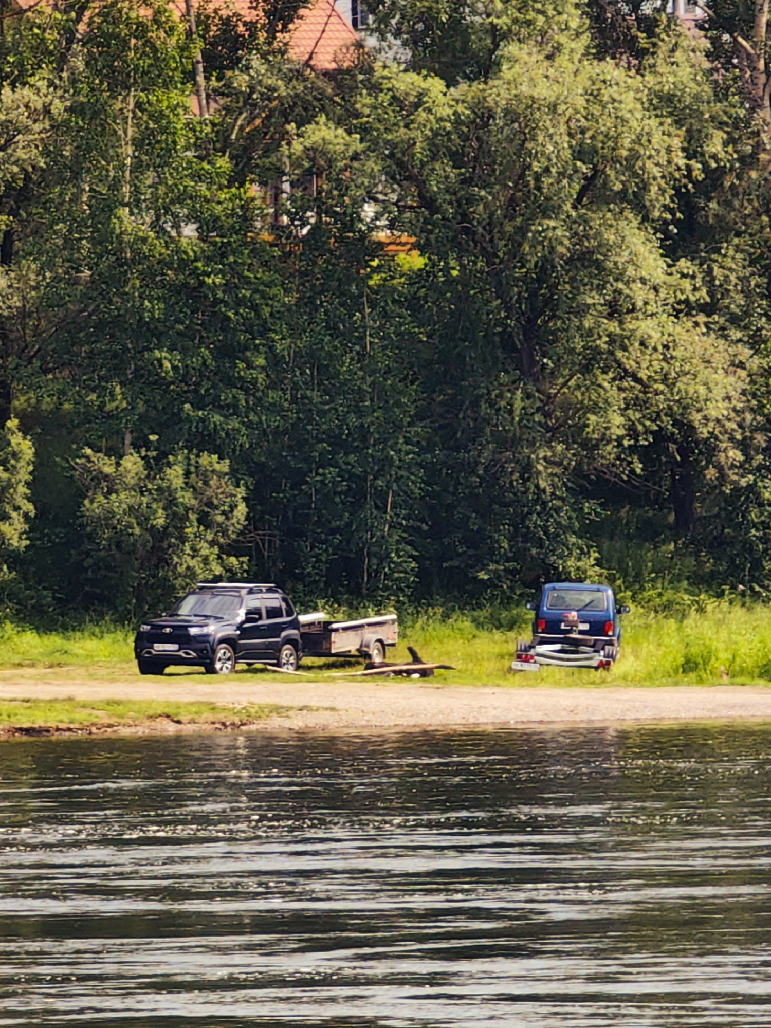 The image depicts a scene by a body of water, likely a river, with a densely wooded background. In the foreground, the water exhibits a ripple effect, suggesting a slight current. On the bank, two vehicles are parked. To the left, a dark-colored SUV is hitched to a trailer. To the right, a blue van is also parked, facing away from the viewer, with a trailer in front of it. The surrounding area is covered in green grass and various types of foliage. Behind the vehicles, a dense line of trees forms a natural barrier. In the upper portion of the image, glimpses of a building with a red roof can be seen through the trees. The overall impression is of a natural setting, possibly a recreational area, located near Sosnovoborsk, Russia.