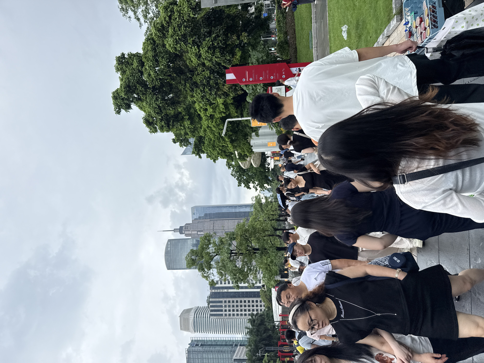A crowd of people walks along a paved path in Tianhe District, China, with a backdrop of modern skyscrapers and lush green trees. The sky is overcast. Many individuals are dressed in casual, everyday clothing, suggesting a public gathering or a busy street. In the foreground, several people are walking away from the camera, their backs visible. To the right, a red signpost with white text and circular icons stands beside the path. A shopping cart filled with small, round containers is visible on the right side of the image, near the edge of the path. The overall scene depicts a lively urban environment.