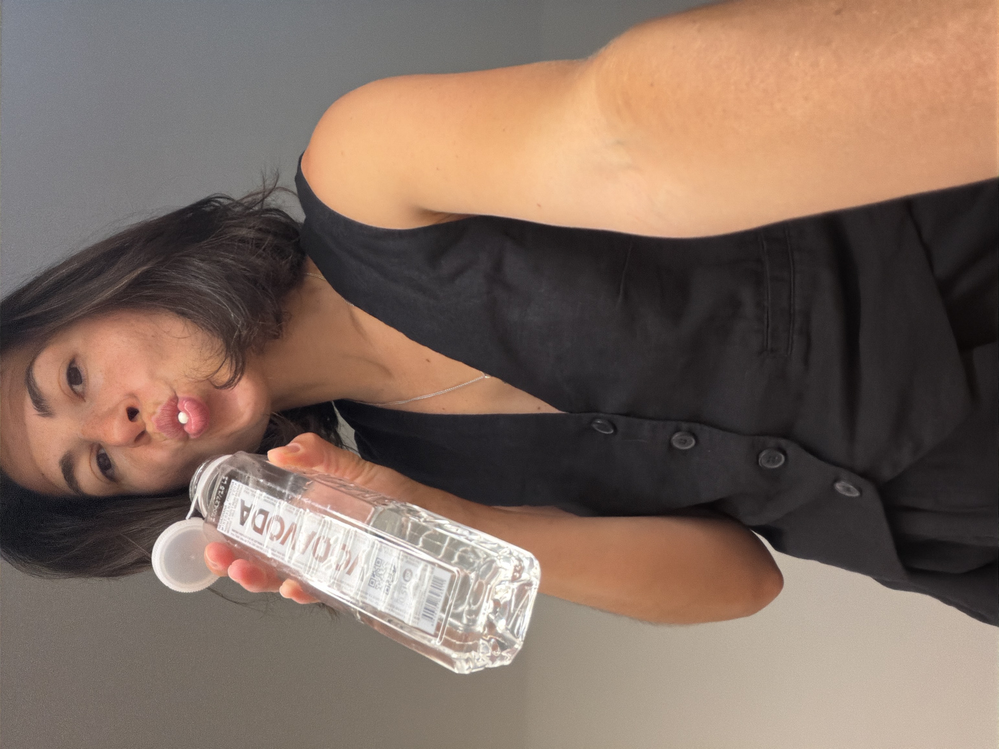 A young woman with dark hair and fair skin is depicted in a close-up selfie, rotated 90 degrees clockwise. She wears a black sleeveless top with visible buttons and a silver necklace. Her expression is playful, characterized by a "duck face" with a small white pill resting on her lower lip. She holds a clear plastic bottle, likely containing water, with a detached white cap in her right hand. The bottle clearly displays "VODA" in reverse text, along with other smaller labels and a QR code. The background is a plain light grey wall, indicating an indoor setting. The overall mood of the image is casual and quirky. This image is contextualized as being from Budva, Montenegro.