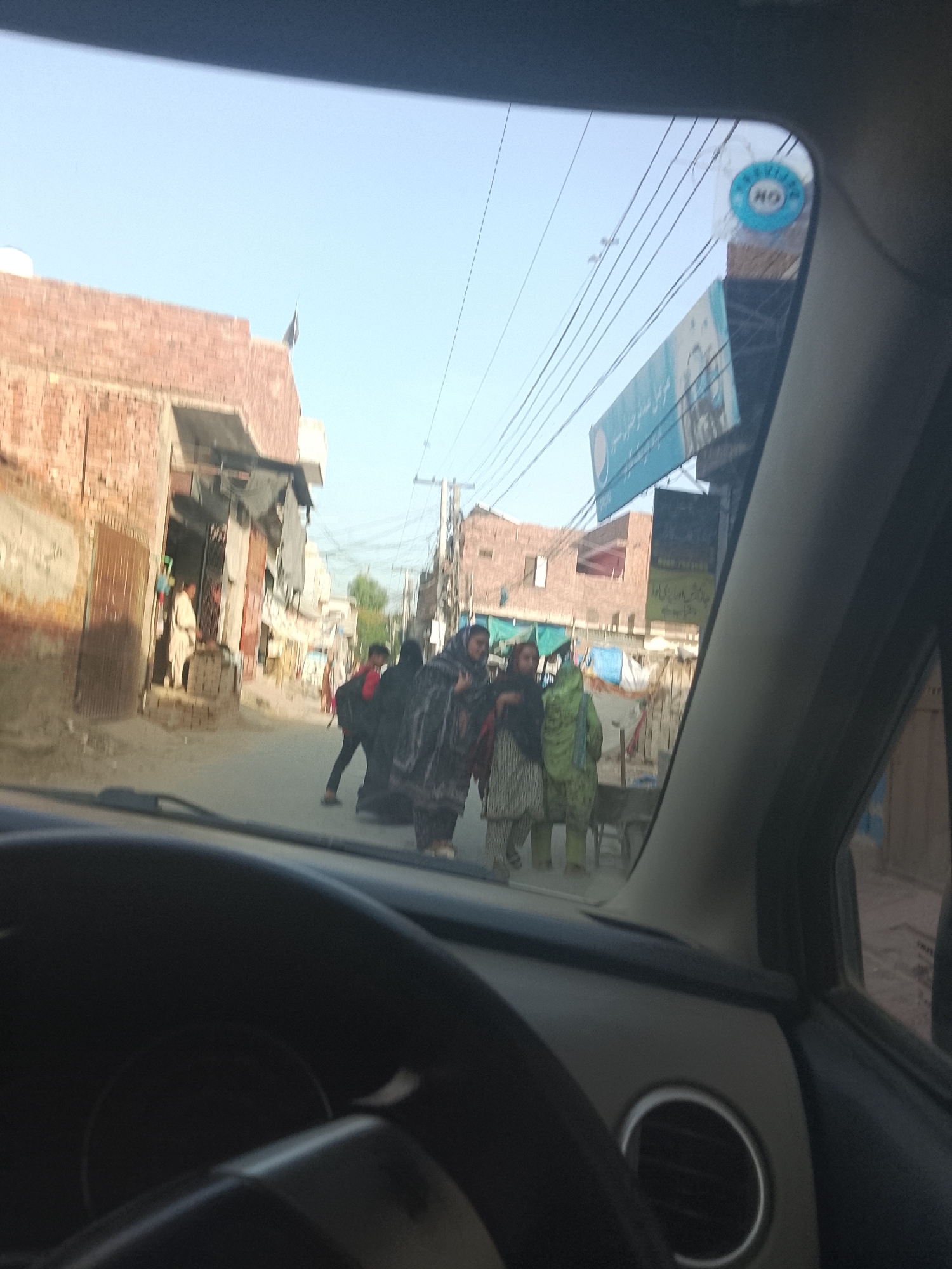 The photo, taken from inside a car, depicts a street scene in what appears to be a South Asian city. Several people are walking in the street. The buildings are brick and appear old. There are power lines overhead. In the foreground, parts of the car's interior, including the dashboard, steering wheel, and air vent, are visible.