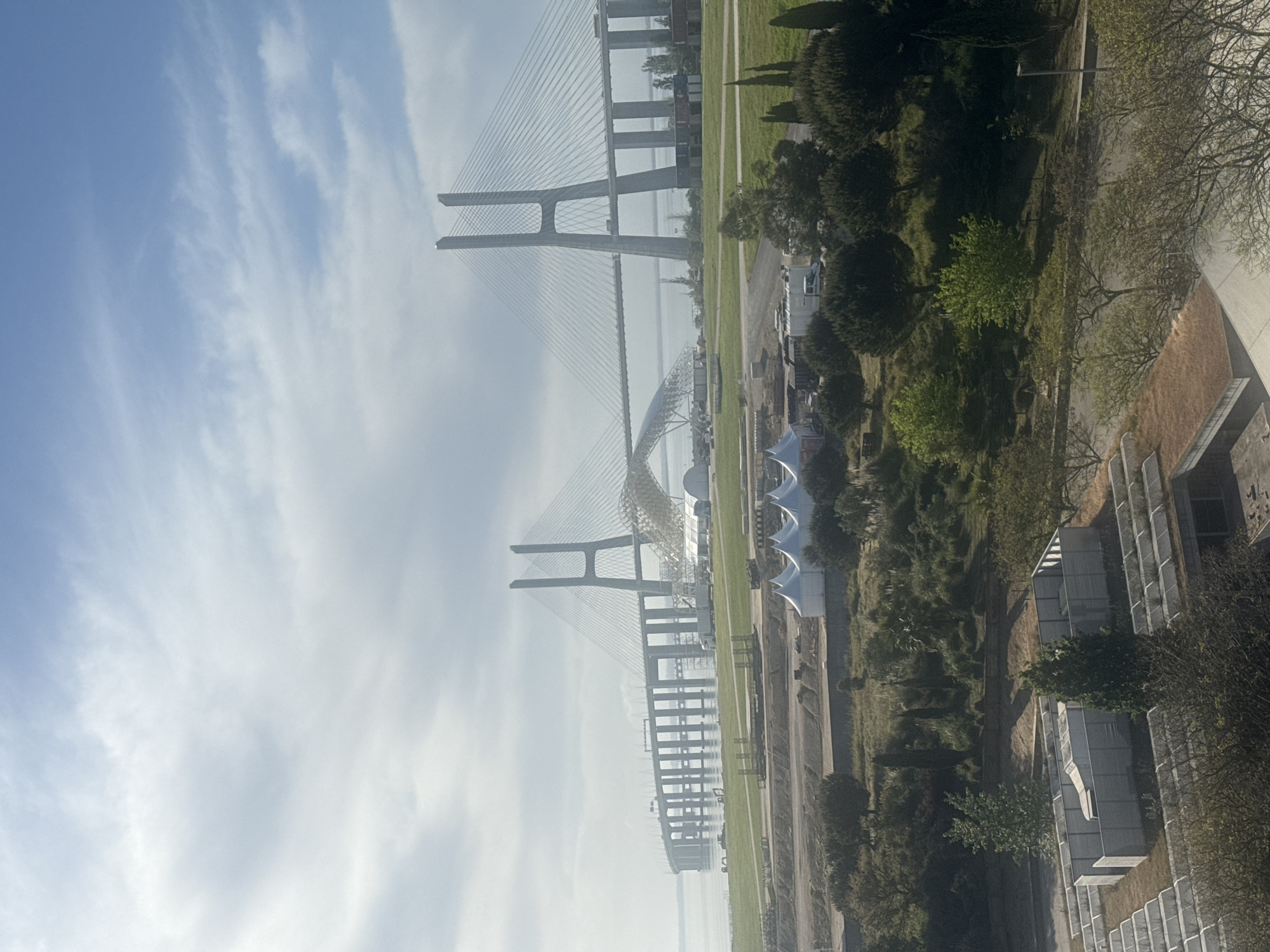 The image depicts a view of the Vasco da Gama Bridge in Loures, Portugal, under a partly cloudy sky. The cable-stayed bridge spans a body of water, with its towers prominently visible. Adjacent to the bridge is a grassy area, interspersed with trees and a series of white tent-like structures. To the right of the frame, the tops of modern buildings are visible amidst greenery.
