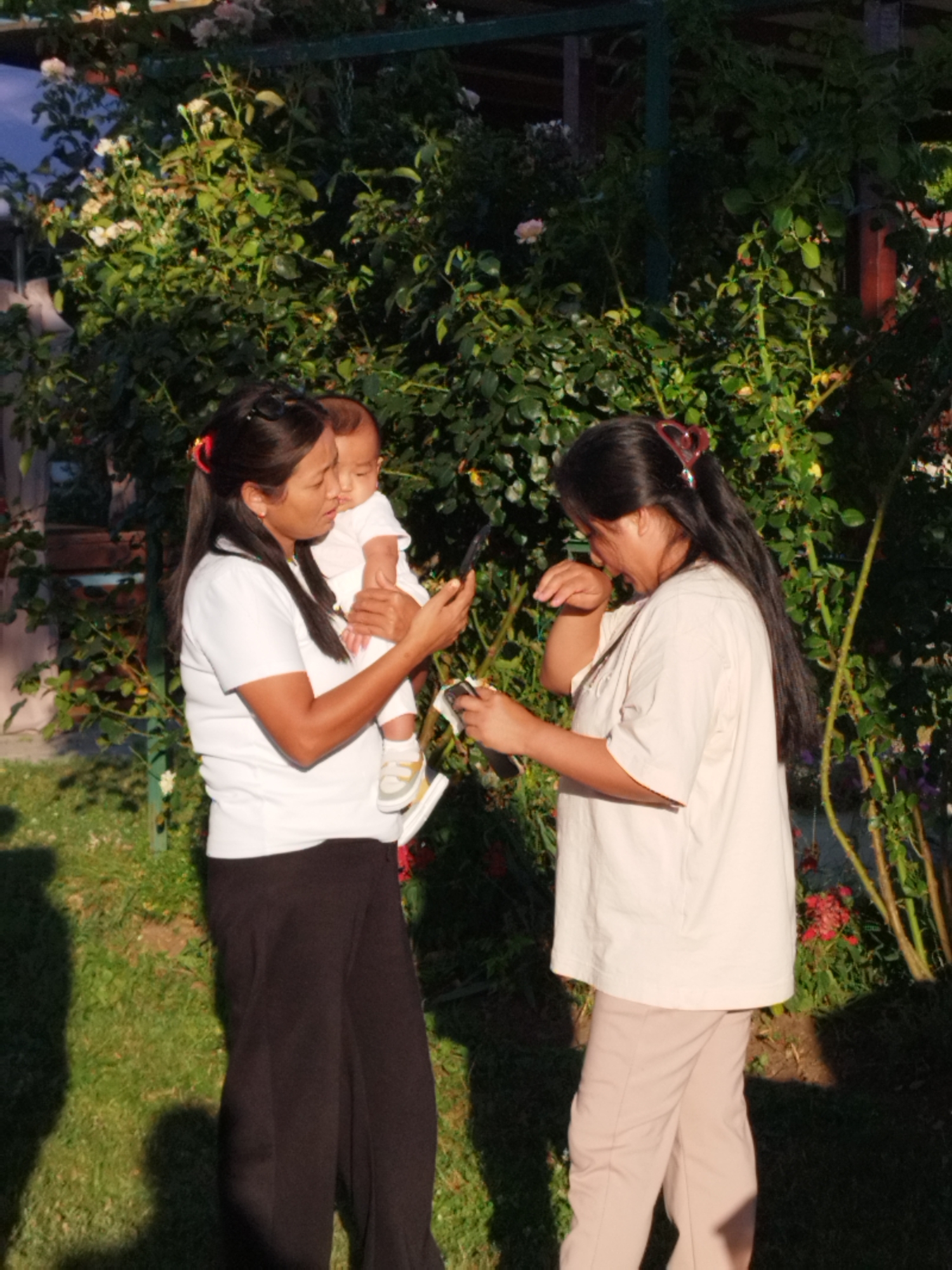 Two women and a baby are outdoors in front of a dense green bush with flowers. One woman on the left is holding a baby and looking at a phone in her hand. She is wearing a white t-shirt and black pants. The baby is wearing a white onesie and yellow shoes. The second woman on the right is wearing a cream-colored t-shirt and tan pants. She is also looking at a phone. The scene appears to be a garden in a residential area. The lighting suggests it is daytime.