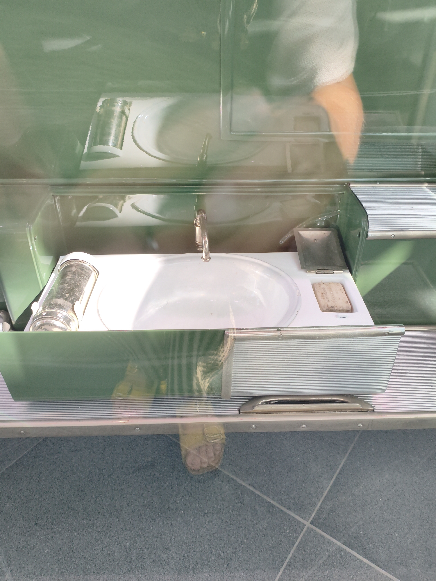 The image shows a vintage sink and soap dish inside a green, possibly retro-styled, vehicle. A chrome dispenser is to the left of the sink. A person's feet, wearing sandals, are visible in the lower part of the frame, suggesting the viewer is standing on a tiled floor. The scene is indoors, possibly in a museum or exhibition space. There is no indication of the city of Ingolstadt, Germany, in the image or the provided context.