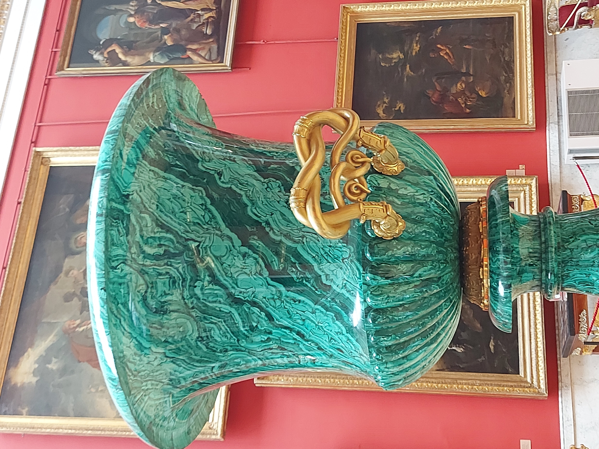 A large, green malachite vase with gold handles sits in front of a red wall adorned with framed paintings. The vase is the main subject, featuring a bulbous body and a fluted pattern. The gold handles are ornate and twisted. The paintings on the wall are in gold frames and appear to depict scenes with figures. The wall is painted red. The location appears to be in a city with many museums.