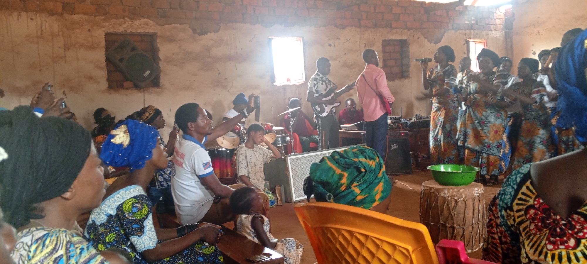 The image depicts a gathering of people inside a brick building in what appears to be Ngalula, Democratic Republic of the Congo. The building, possibly a church, features unfinished brick walls with multiple windows. A group of people, primarily women, stand together, singing and clapping, while a band with instruments including guitars and drums plays. In the foreground, several people sit and observe the scene, some holding phones. A green bowl sits atop a drum. A loudspeaker is wedged into a window opening.