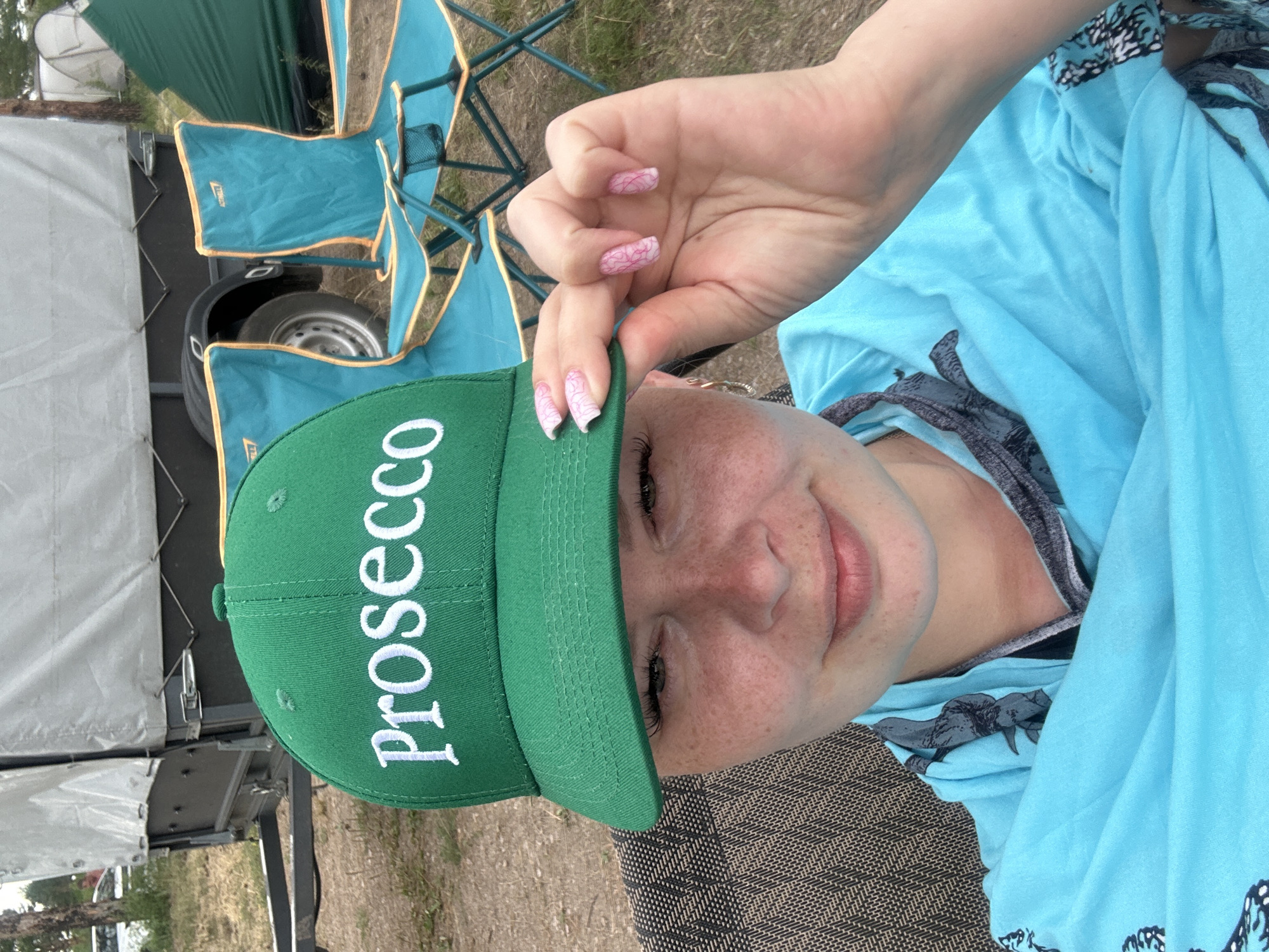 The image shows a close-up, low-angle shot of a young woman outdoors, wearing a bright green baseball cap with white lettering that spells out "Prosecco." She has fair skin with freckles and is wearing a blue hooded shirt. Her right hand, with pink nail art, is holding the brim of the cap. The background is blurred but shows elements of an outdoor setting, including what appear to be camping chairs, a trailer, and vegetation. The lighting suggests it is daytime, possibly sunny. The woman's expression is relaxed and happy. The word "Prosecco" is clearly visible on the cap. The context of the image is related to an outdoor gathering or activity, possibly a picnic or camping trip.