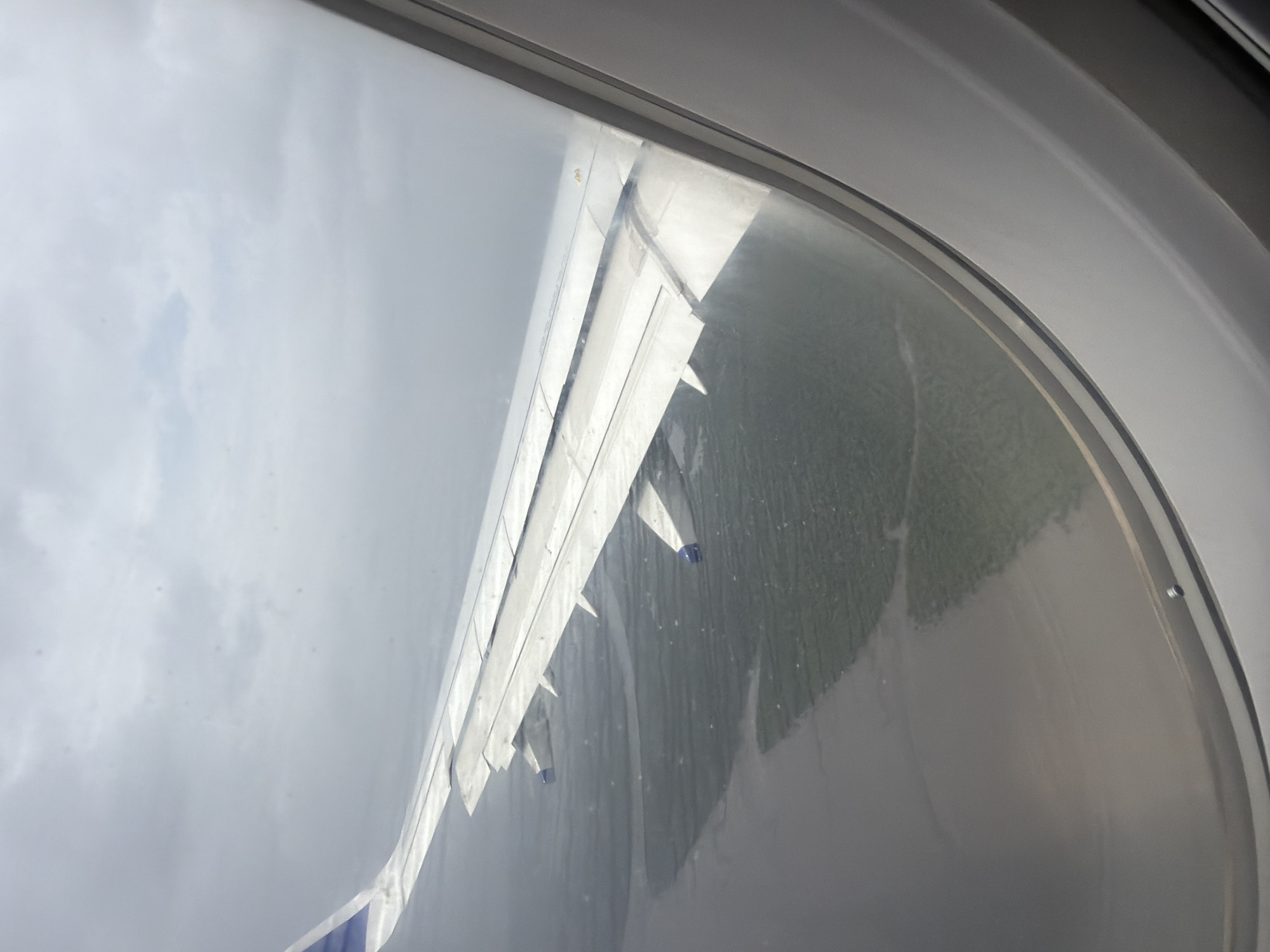 An aerial view from an airplane window shows the wing of the aircraft extended to the left. The wing is white with blue accents at the tip. Below the wing, a body of water, possibly a bay or inlet, meets land covered in green vegetation. The water appears somewhat muddy or murky. The sky above is overcast with thick gray clouds. The image is framed by the airplane's window, with the white interior of the window frame visible on the right side of the picture. The scene is likely over the coastal region near Vakrul, India, given the appearance of water and vegetation.