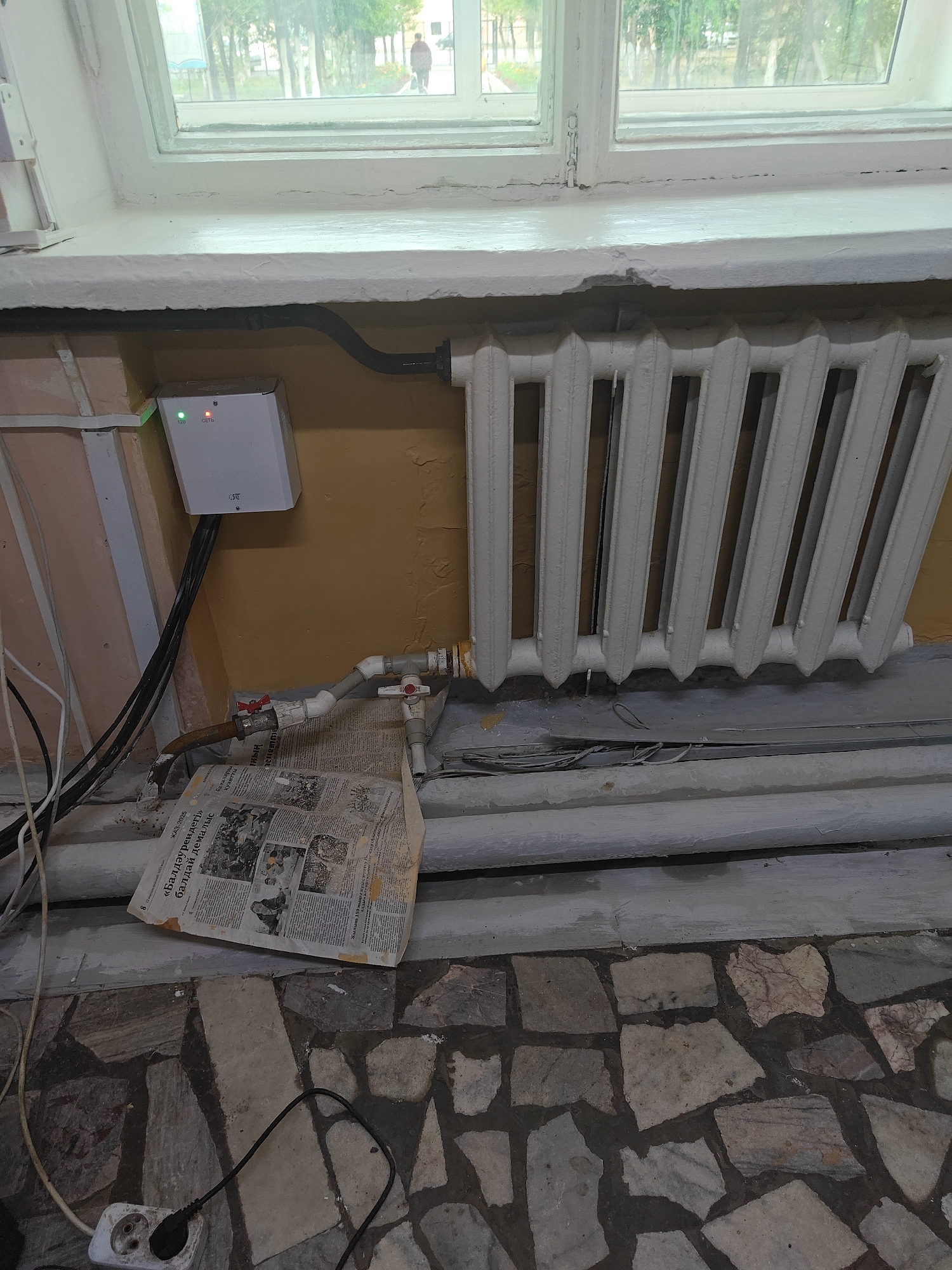 A white electronic device with green and red indicator lights is mounted on a beige wall next to a white cast-iron radiator. The device has "128" and "СЕТЬ" written below the lights. Black cables descend from the device. Below the device, a newspaper is spread out, partially covering pipes. A red valve is visible on a pipe connected to the radiator. The scene appears to be indoors, with a window in the background showing trees and a person walking outside. The floor is made of irregular stone tiles. A white electrical outlet with a black plug is visible in the lower left corner. The context of Ekibastuz, Kazakhstan, is not directly evident from the visual elements of the image.