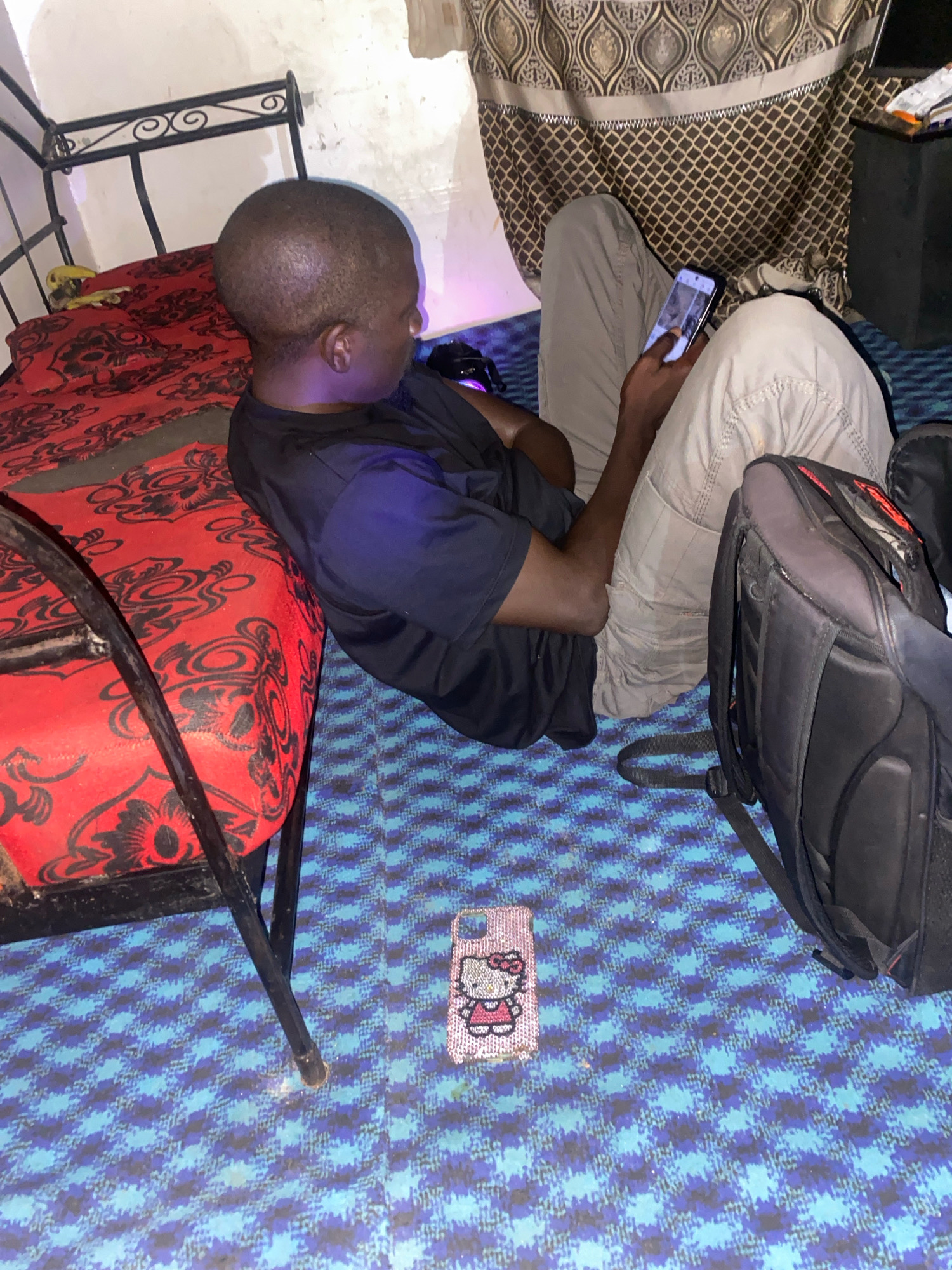 The image depicts an indoor scene, likely a bedroom, situated in Jos, Nigeria. A young Black male is seated on the floor, leaning against a bed with a red and black patterned mattress. He is wearing a black t-shirt and light-colored pants, and is holding a smartphone in his right hand, looking at the screen. To his left, a black backpack rests on the floor. Nearby, on the blue patterned carpet, a phone case adorned with a Hello Kitty design is visible. The room appears dimly lit, with a metal-framed bed and patterned curtains in the background. The overall impression is one of a casual, personal space.