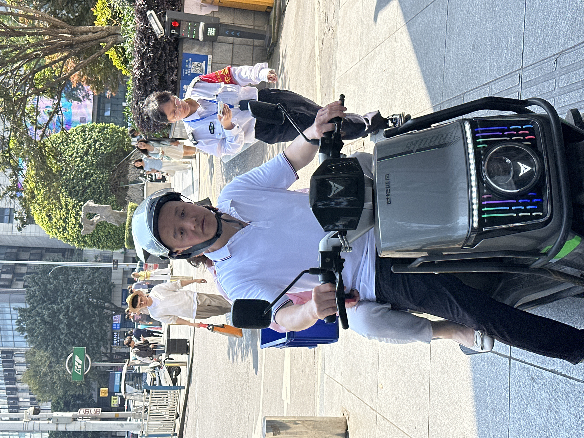 A man wearing a light blue helmet and white polo shirt is seated on a gray electric scooter, facing the camera. The scooter has a distinct head unit with colorful LED lights. The location is Yuzhong District, China, discernible by the presence of Chinese characters on nearby signage and architectural elements. The scene is a bustling urban area, evidenced by the presence of pedestrians, trees, and buildings in the background. Another man wearing a white uniform with a red armband is walking parallel to the scooter, slightly behind. Other pedestrians are walking on the sidewalk in the background.