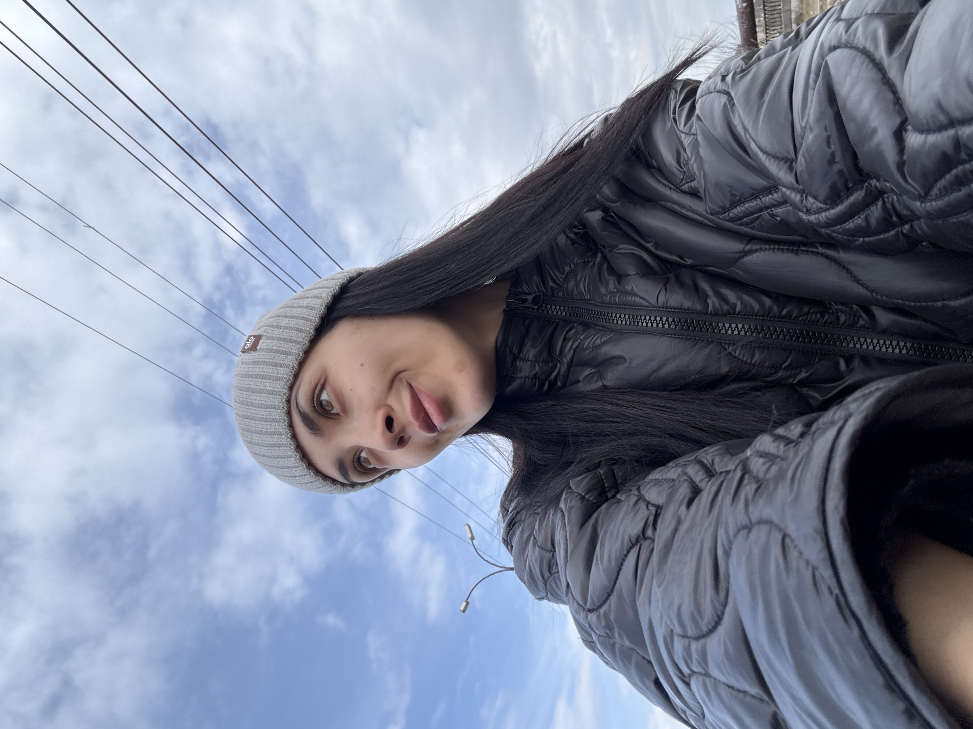 A young woman is taking a selfie outdoors on what appears to be a partly cloudy day. She is wearing a gray beanie and a black quilted jacket. Electrical wires are visible above her, and streetlights are behind her. The sky is light blue with white clouds. The location is Votkinsk, Russia.