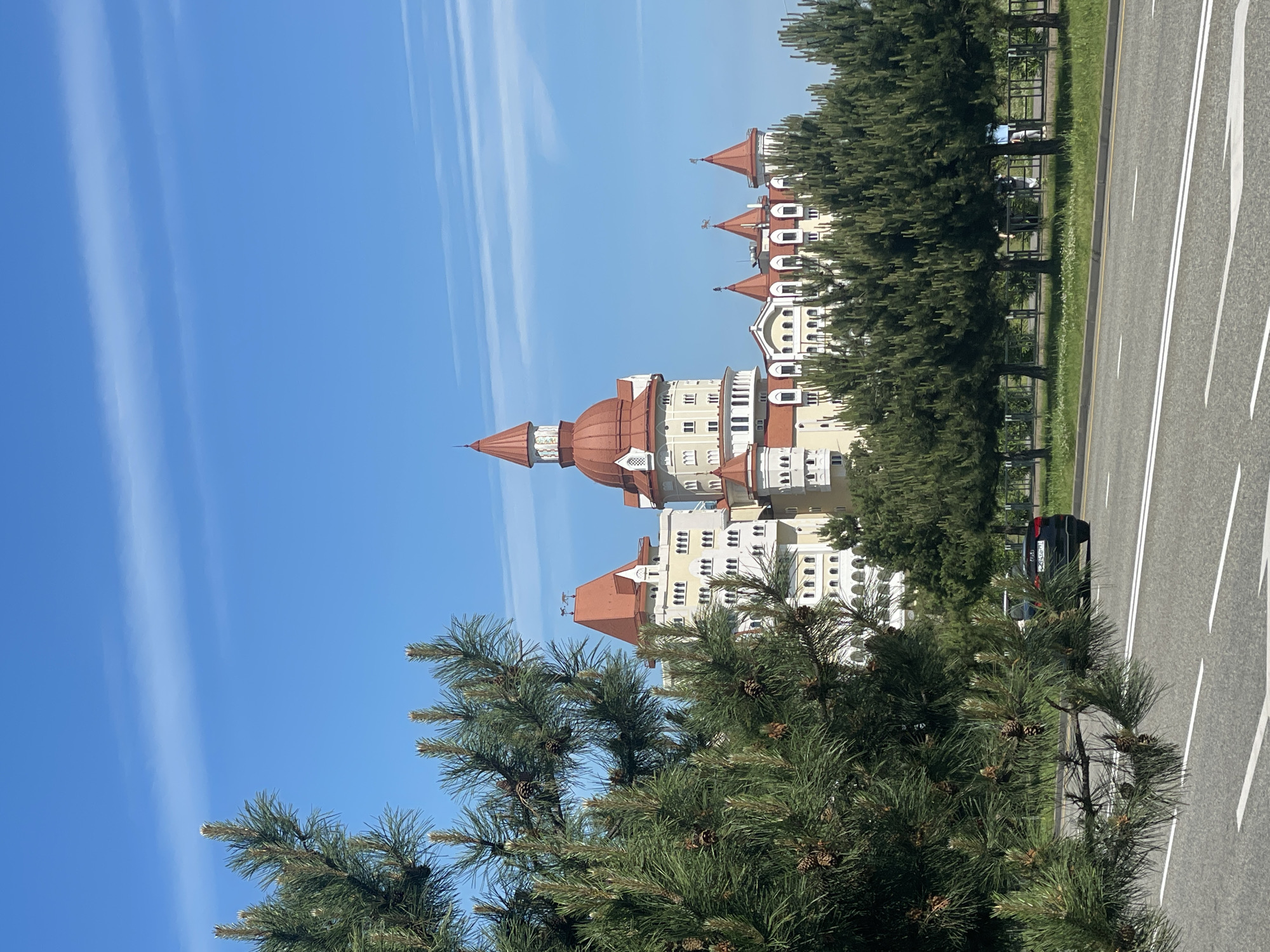 The image shows a tall building with red roofs, resembling a castle, located in the city of Sirius, Russia. The building is light-colored with multiple windows and turrets. A pine tree with cones is visible in the foreground, partially obscuring the view of the building. To the right, there is a road with white lane markings, and a black car is parked on the side of the road. Additional trees line the road. The sky is blue with light streaks, suggesting either clouds or contrails.