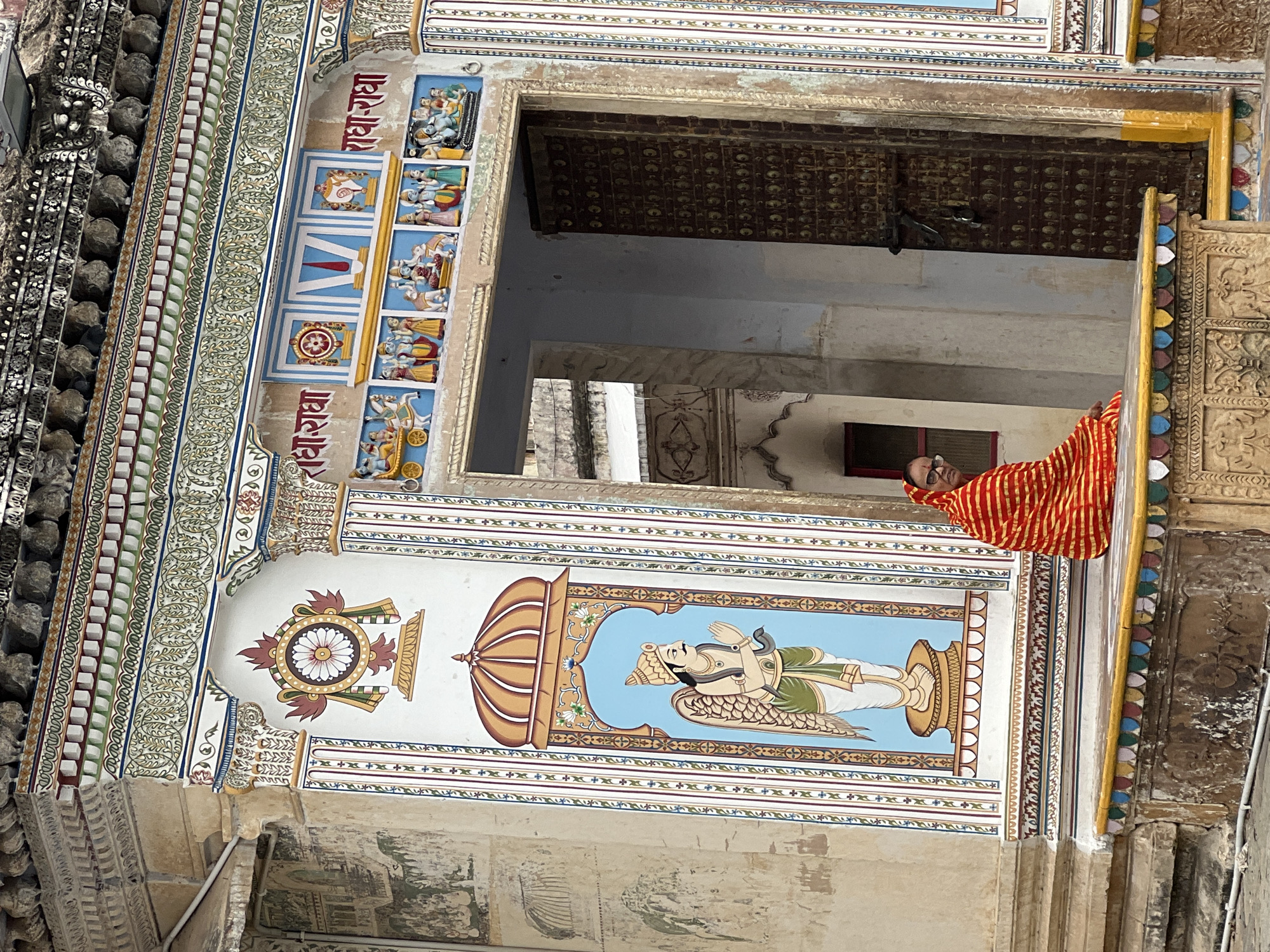 This is a medium-shot, eye-level photograph of a woman sitting on a decorative ledge in Pushkar, India, in front of a vibrantly painted building. The building exhibits traditional Indian architectural and artistic styles, with intricate carvings, colorful murals, and ornate detailing. The woman is wearing a red and orange striped sari and glasses. She is looking towards the camera. To her left is a mural of a winged figure with hands clasped in prayer. Above the murals are inscriptions in Hindi. To her right is a large, weathered wooden door. The ledge she is sitting on is adorned with colorful patterned tiles. The background shows more of the building's facade, including more painted figures and decorative elements. The lighting suggests it is daytime.