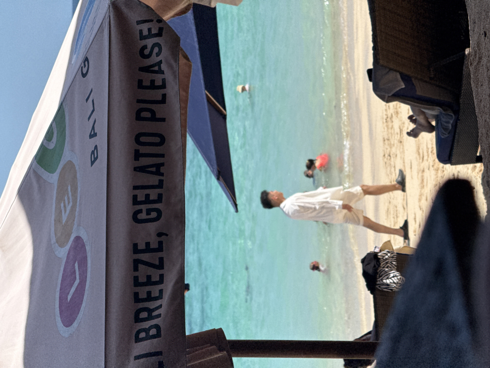 The image depicts a beach scene in Kuta Selatan, Indonesia, from a slightly obscured perspective. An advertisement for "Bali Breeze, Gelato Please!" is visible on an umbrella in the foreground. The turquoise water meets a sandy beach where several people are present. A man in a white shirt and shorts is walking along the shoreline. Others appear to be swimming or wading in the water. Beach chairs are situated on the sand.