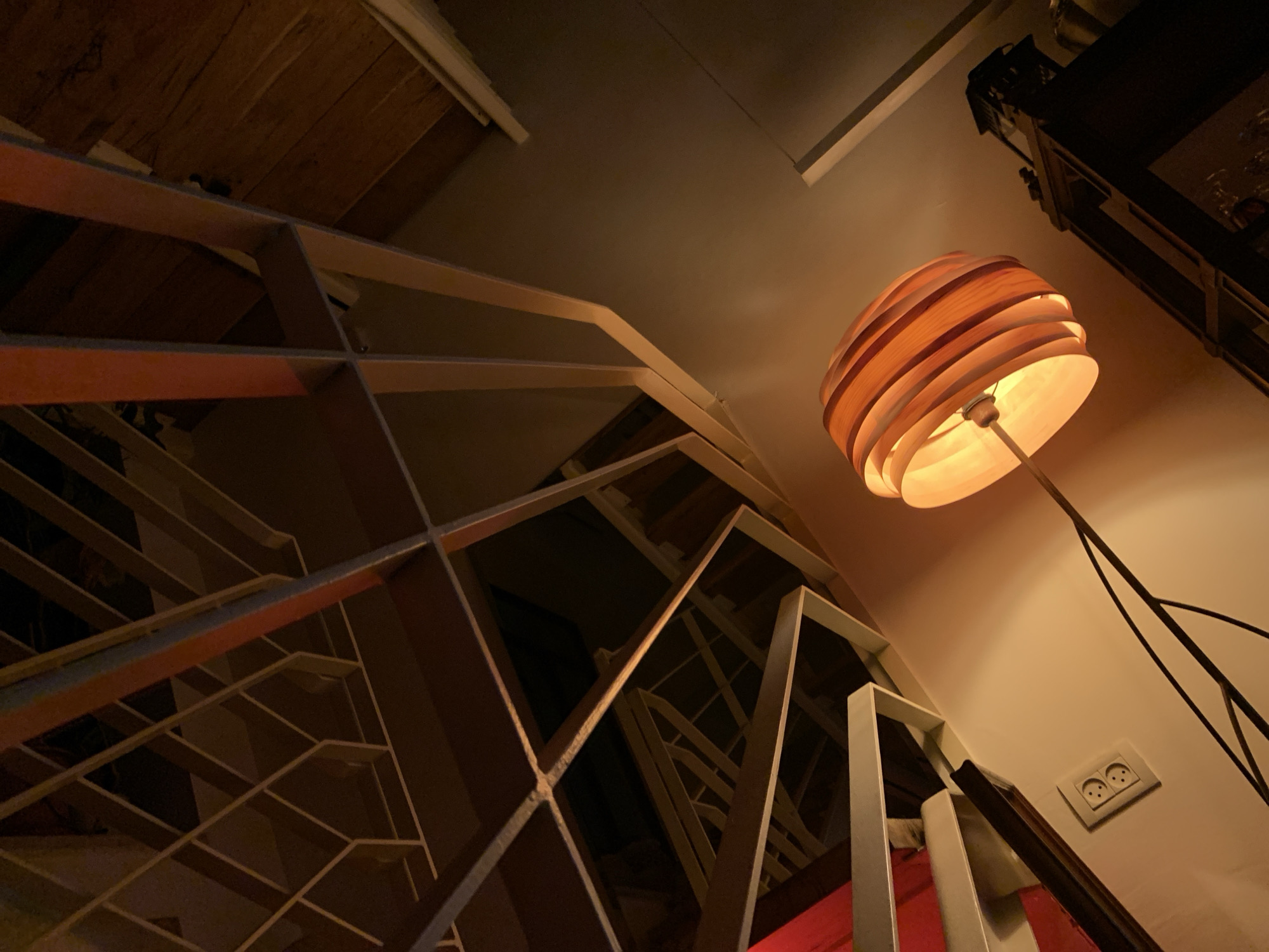 A low-angle shot in Tel-Aviv, Israel, reveals an interior space with geometric stairs and a wooden floor lamp. The light, composed of stacked wooden pieces, casts a warm glow on the adjacent white wall, near a standard wall socket. The stairs, constructed with thin white metal frames, ascend in an orderly zigzag. The background includes a wooden ceiling and a dark cabinet.