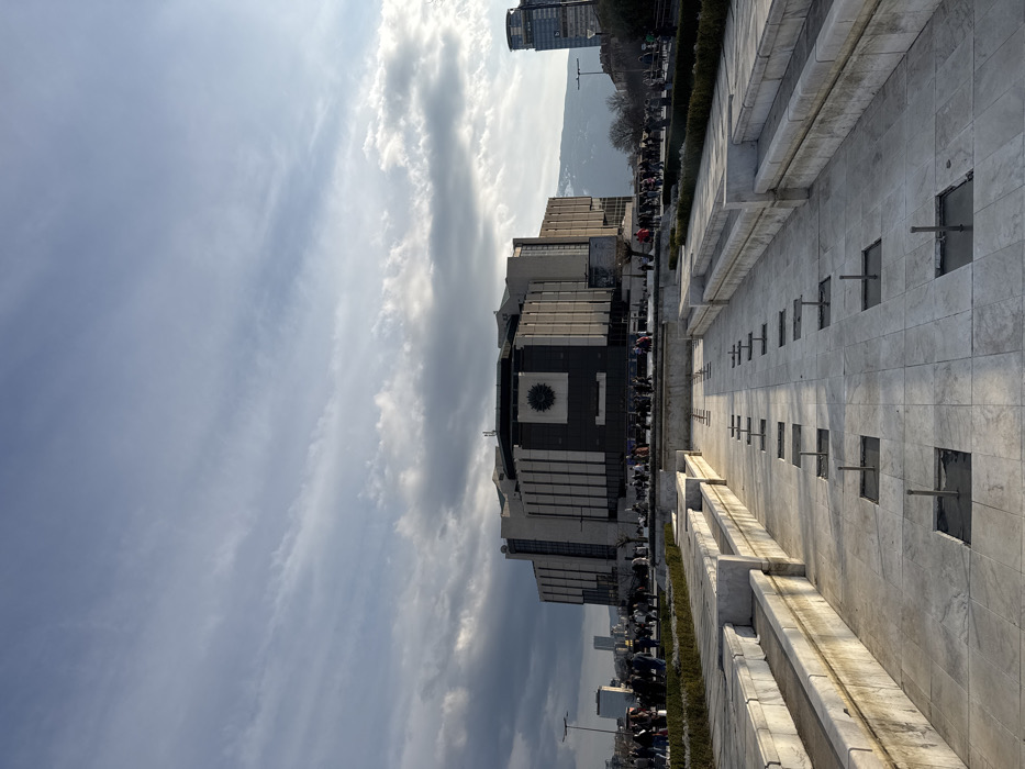 The image displays the National Palace of Culture (NDK) in Sofia, Bulgaria, viewed from a slightly elevated perspective across its forecourt. The NDK is a large, multi-story Brutalist-style building with a distinctive facade featuring numerous vertical concrete fins and a prominent central square panel containing a clock-like emblem. In the foreground, a wide, multi-tiered fountain structure, constructed from light-colored stone or marble, occupies the lower half of the frame. The fountain is currently dry, with multiple square basins containing exposed metal piping elements. Numerous individuals are visible as small figures in the mid-ground, standing and walking on the paved plaza directly in front of and to the sides of the NDK. The sky is partially overcast with a mixture of high, wispy clouds and lower, denser cloud formations. In the distant background to the right of the NDK, snow-capped mountains are visible. Additional urban high-rise buildings are discernible in the background on both the left and right sides of the main structure. Low-lying evergreen landscaping flanks the edges of the dry fountain complex.