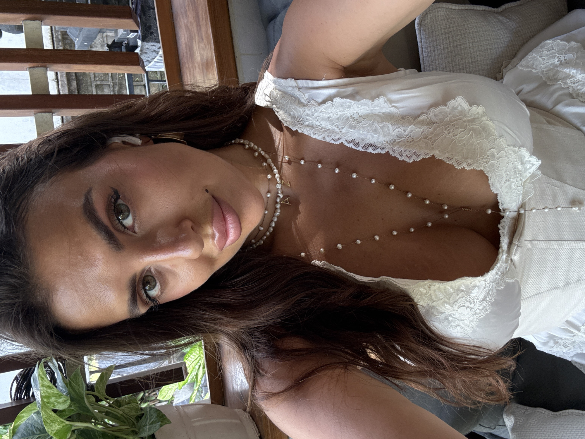 A tan-skinned woman with dark hair and green eyes is captured in a close-up photograph. She is wearing a white, low-cut top with lace detailing and a pearl necklace. The background reveals a partial view of wooden stairs, green foliage, and a glimpse of what appears to be urban scenery. Based on the prompt's context, the location is North Kuta, Indonesia.
