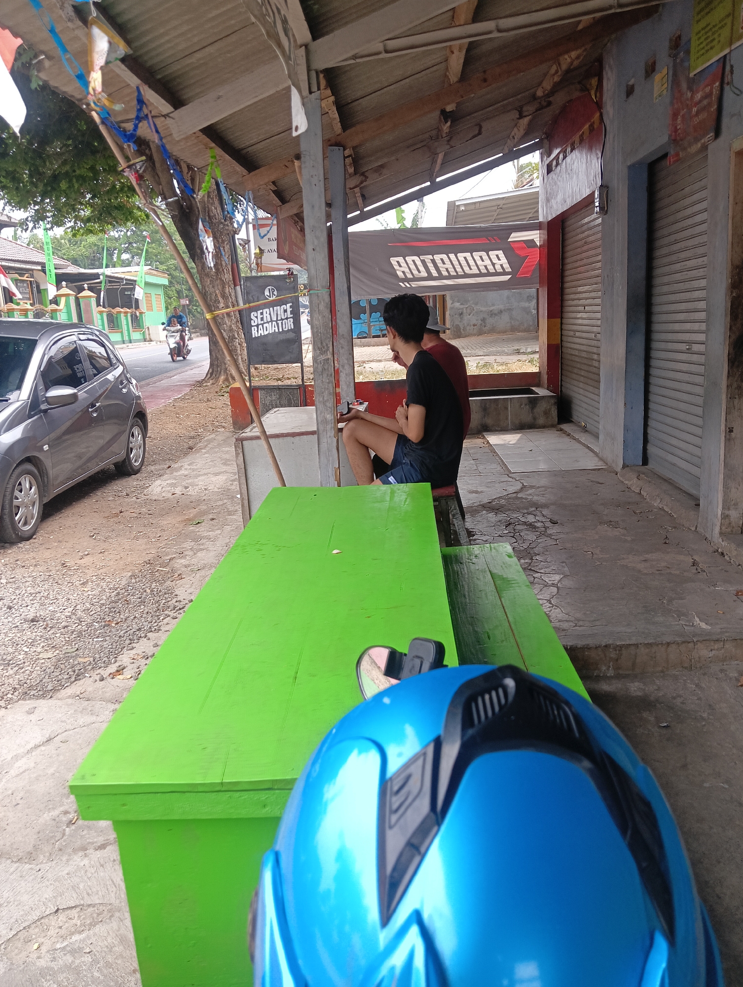 An outdoor scene captures a street-side establishment in Manggari, Indonesia, with a focus on a bright green wooden picnic table in the foreground. The table is set on a concrete and gravel surface, adjacent to a gray car parked on the left. Behind the table, two individuals are seated on a bench under a corrugated metal awning supported by wooden posts. One person, wearing a black t-shirt and blue shorts, faces away from the camera, while the other, in a maroon shirt, is partially visible. A dark blue motorcycle helmet rests on the table. Further in the background, a street stretches out with a person on a motorcycle and a building with rolled-down metal shutters. A sign reading "SERVICE RADIATOR" is visible on a pole near the awning. Decorative banners and wires are strung across the top of the awning.