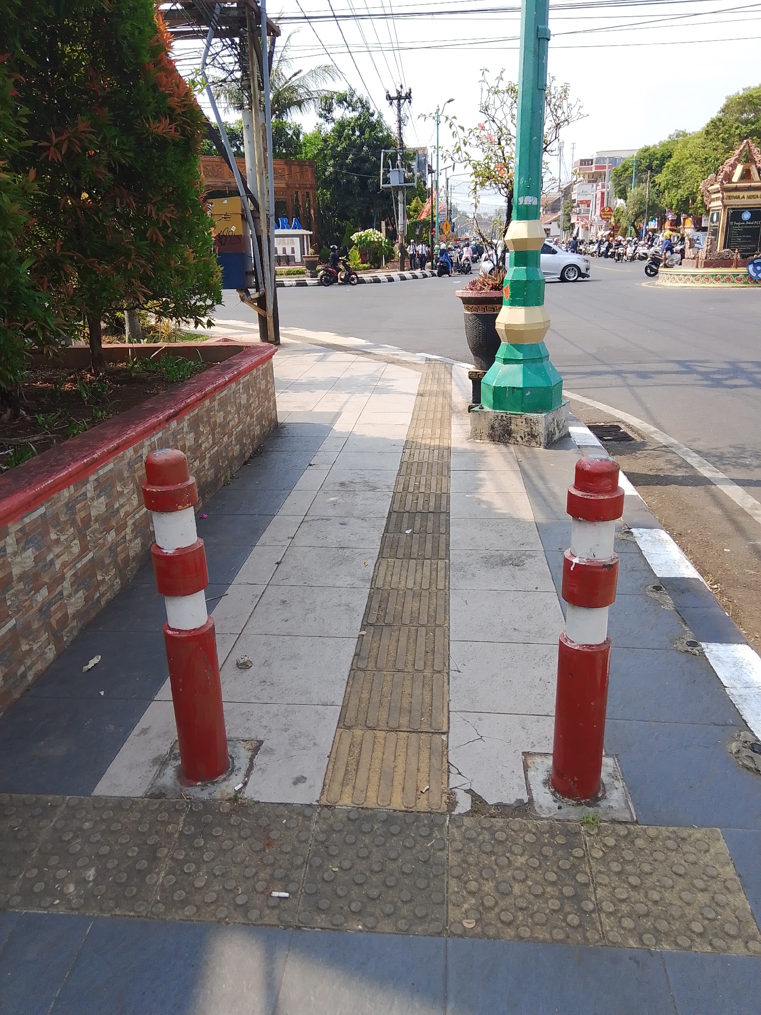 The image shows a street scene in Jepara, Indonesia, likely near a public monument or intersection. In the foreground, two red and white striped bollards stand on a sidewalk. The sidewalk features tactile paving for the visually impaired. A low, brick wall with a red trim borders a planter box with a tree. In the background, a street with moderate traffic is visible, including cars and numerous motorcycles. A distinctive green and gold lamppost stands prominently near the sidewalk. Buildings and other street furniture are present, and the sky is clear and bright. The overall impression is of a typical day in a bustling Indonesian city.