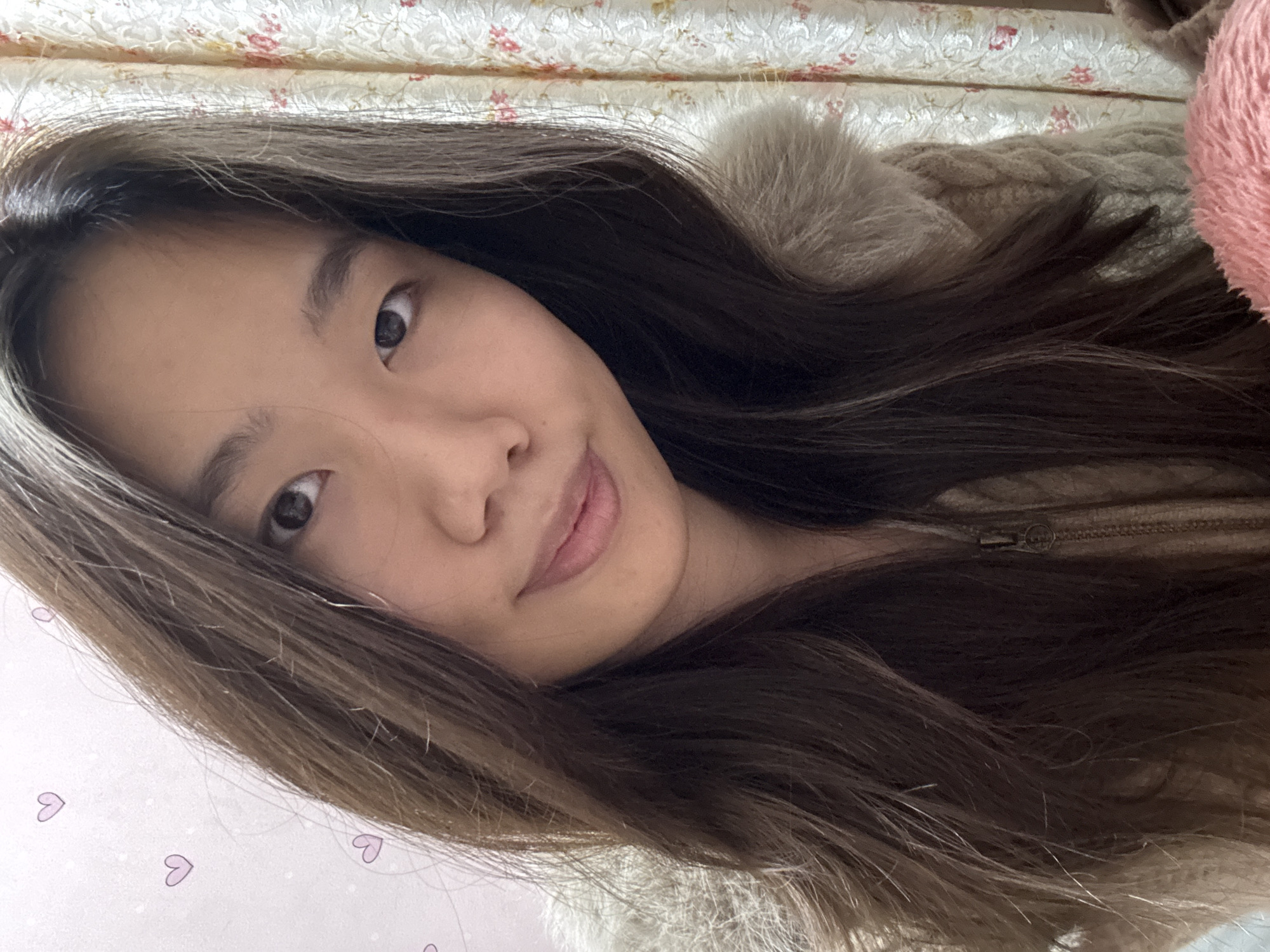 The image depicts an indoor scene in Shapingba District, China, featuring a female individual of East Asian descent. The subject is positioned facing slightly right of center, looking directly at the camera with a subtle closed-mouth smile. She has long, dark brown hair parted on the left side, which cascades over her shoulders. She is wearing a light beige or tan knitted garment, possibly a cardigan or sweater, featuring a visible zipper pull on the front and a faux fur trim around the collar or hood.
The background on the left consists of a light-colored wall adorned with small, light purple heart-shaped decals. On the right, a cream-colored curtain with a repeating floral pattern in red and green hues is visible. A blurred, pink, fluffy object is partially visible in the lower right foreground. The lighting is soft and even.