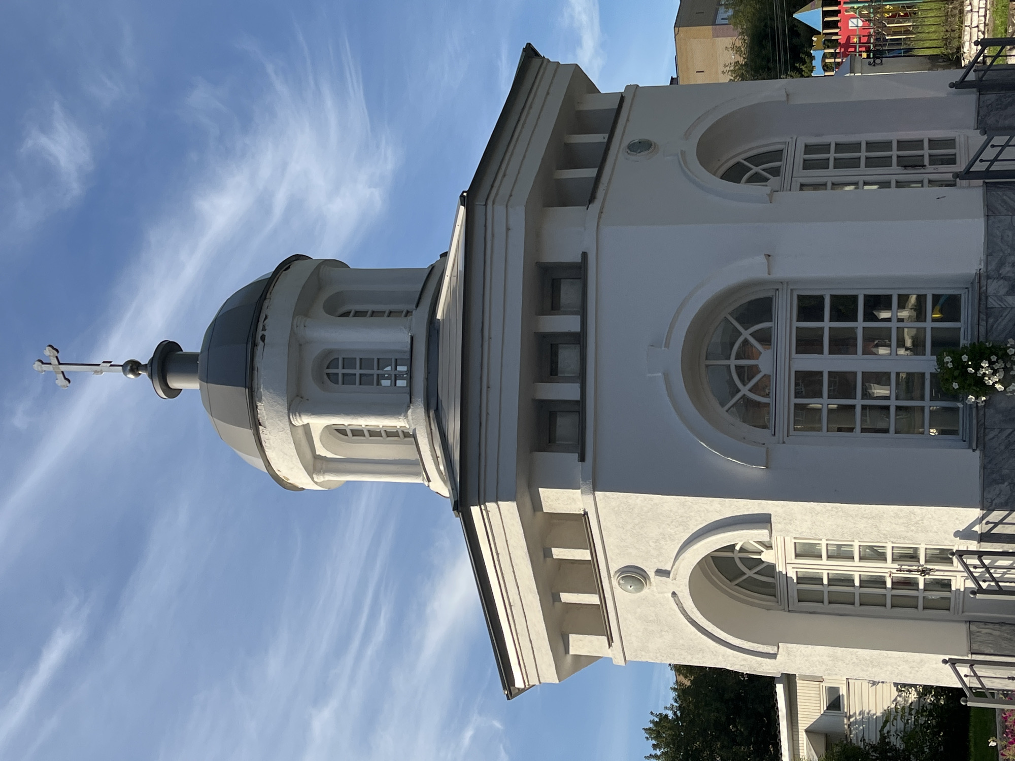 The image shows the exterior of a white, octagonal chapel with a grey dome topped by a cross. The chapel has arched windows with decorative tracery and white window frames, and a white door with multiple glass panes. The building is seen from a low angle against a bright blue sky with wispy white clouds. In the background, there are trees, a smaller building, and a colorful children's playground. This chapel is likely located in Nizhny Novgorod, Russia, given the architectural style often found in Russian Orthodox chapels. There are no people visible in the image. The overall impression is of a bright, clear day with the chapel as the prominent subject.