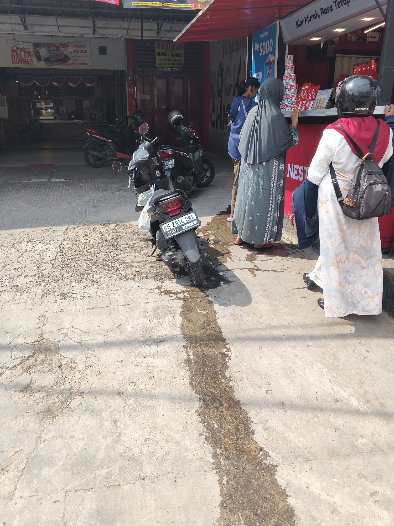 In Bandar Lampung, Indonesia, a street scene unfolds with several individuals interacting at a food stall. The stall, identifiable by a red awning and signage, appears to be a fast-food establishment, possibly selling chicken. A prominent banner advertises "Chicken Steak Murmer" with a price of Rp 12,000. Another sign above the counter reads "BiAR MURAH, RASA TETAP."
Three people are visible at the counter. The person closest to the viewer, wearing a white and light blue patterned dress and a black helmet, has a black backpack. To their left, a person in a dark grey hijab and a patterned dress is engaged with the stall. Further left, partially obscured, is another person in a blue jacket.
Several motorcycles are parked on the paved ground, indicating a mode of transportation common in the area. One motorcycle in the foreground displays the license plate "BE 2914 DBI." The ground is a mix of concrete and rougher textured pavement, with some damp or wet patches. The overall environment suggests an urban setting with commercial activity.