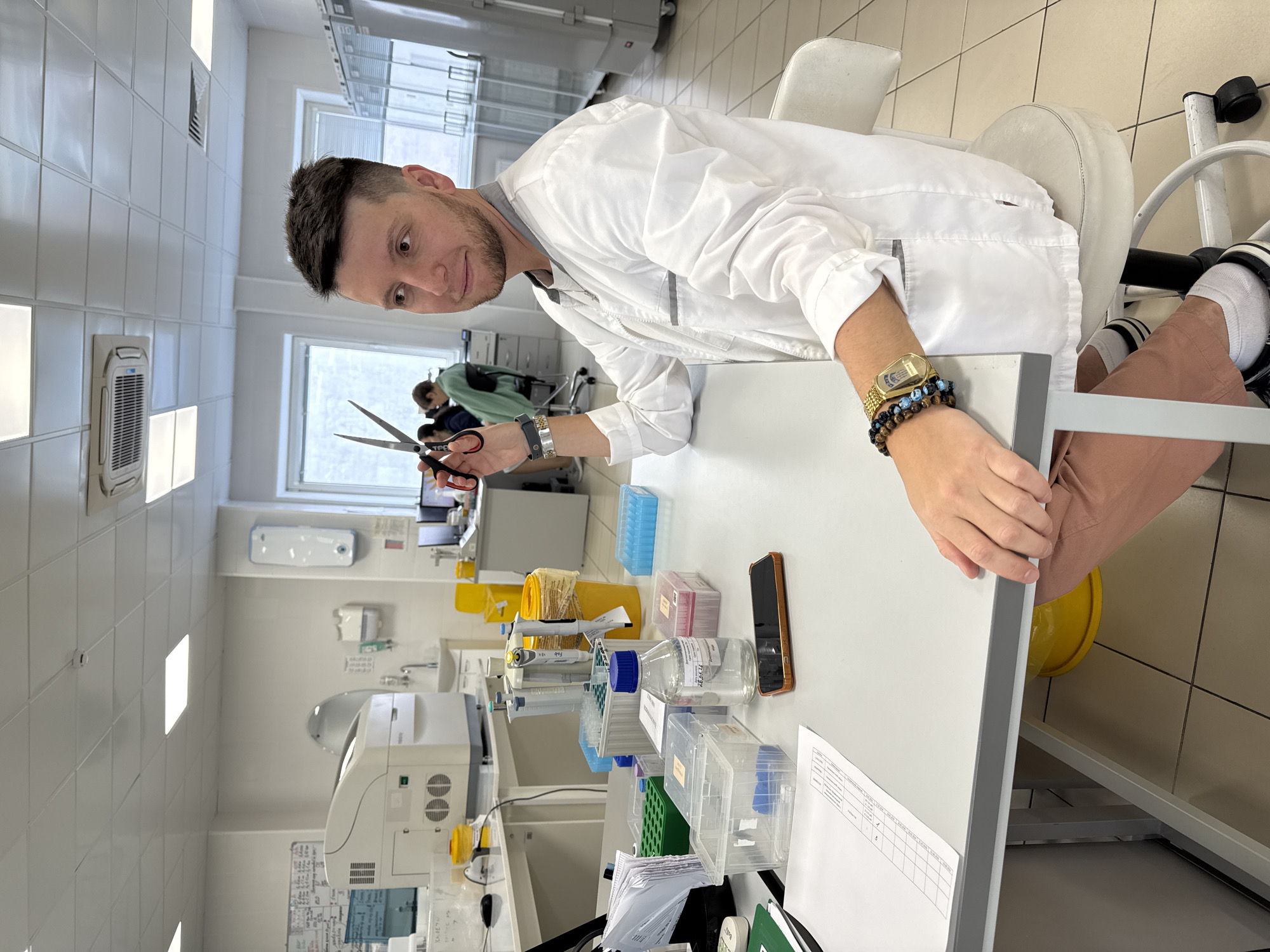 A man in a white lab coat sits at a white lab table in what appears to be a laboratory. He holds scissors in his right hand and looks towards the camera. The table contains a variety of lab equipment and supplies, including disposable pipettes, various test tubes, containers, a mobile phone, and a document. A white analyzer is in the lower left portion of the image. There is a window in the background and others can be seen working in the lab. The man is wearing a gold watch and multiple bracelets on his left wrist. The location is presumed to be Moscow, Russia, based on the user’s specification.