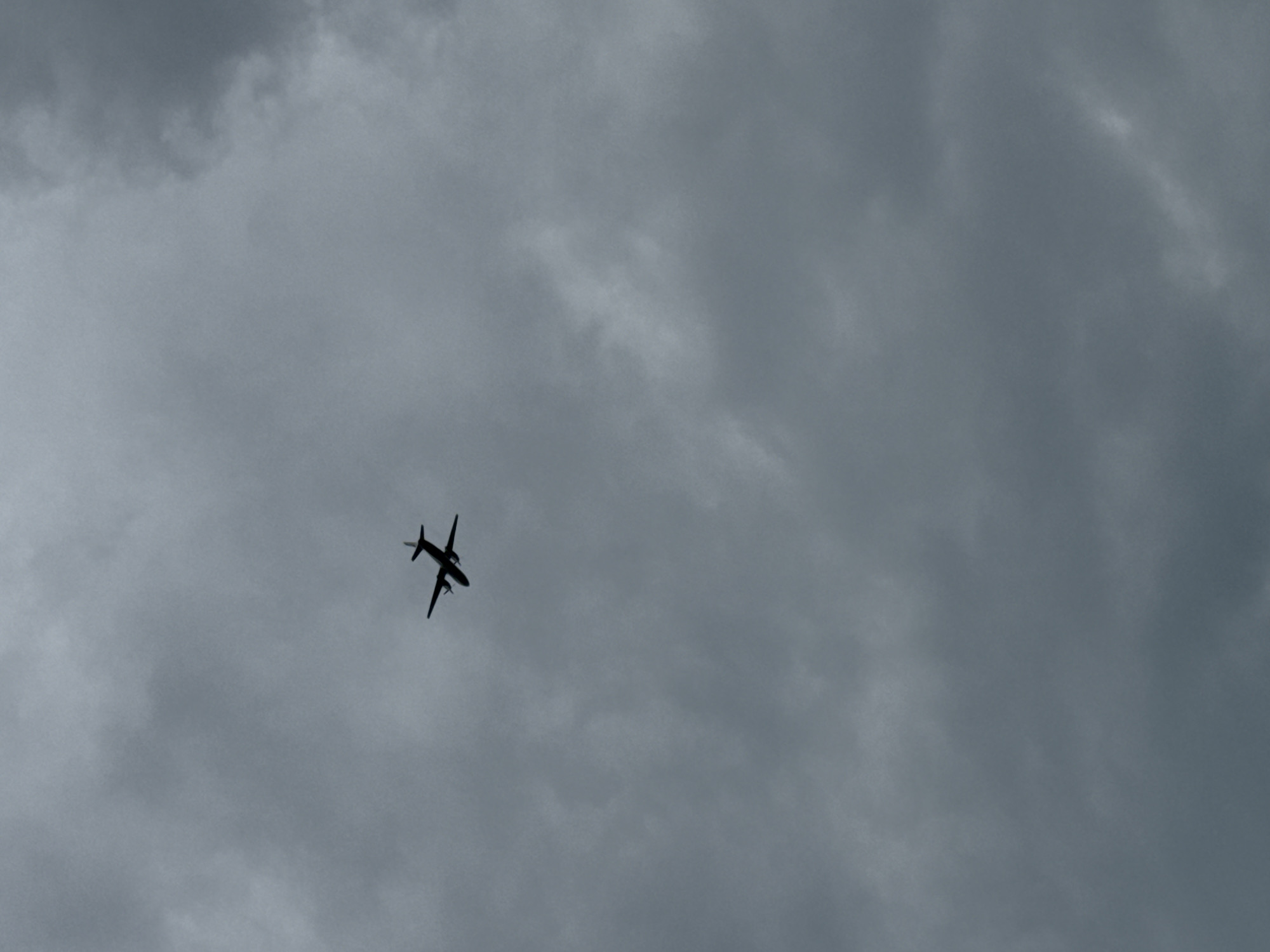 A dark, silhouetted airplane is flying in the sky against a backdrop of grey, overcast clouds. The scene appears to be located in Sirius, Russia. No people or other objects are visible. The plane is in motion.