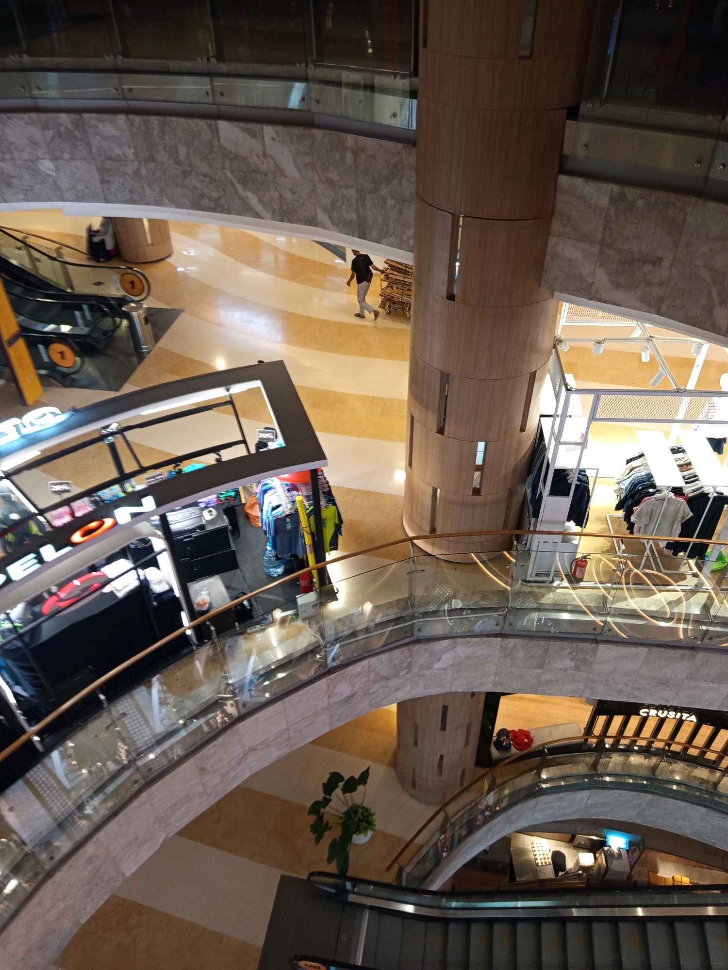 This image depicts an interior view of a multi-level shopping mall in South Jakarta, Indonesia. The perspective is from an upper level looking down onto escalators, walkways, and retail kiosks.
In the foreground, a curved glass and wood railing overlooks a lower level. The floor of the mall features a patterned design of beige and light brown rectangular tiles.
To the left, a retail kiosk is visible with clothing items displayed. A sign with orange illuminated lettering is partially visible. Further back and to the left, an escalator is descending, with a person in a black shirt and light-colored pants walking away from it towards a brown cylindrical pillar. The person is pushing a trolley with what appears to be boxes or merchandise.
The central and right side of the image shows a large, cylindrical wooden pillar structure. Adjacent to this pillar is another retail display area with clothing on racks. A red fire extinguisher is mounted on the wall near this area.
Further down and to the right, another escalator is visible, along with a storefront bearing the name "CRUSITA". Below this, another retail area is visible with lights and merchandise.
The overall environment is well-lit, suggesting a commercial space. The architecture incorporates marble-like stone on the walls and curved glass elements. A potted plant is situated on a lower level walkway.