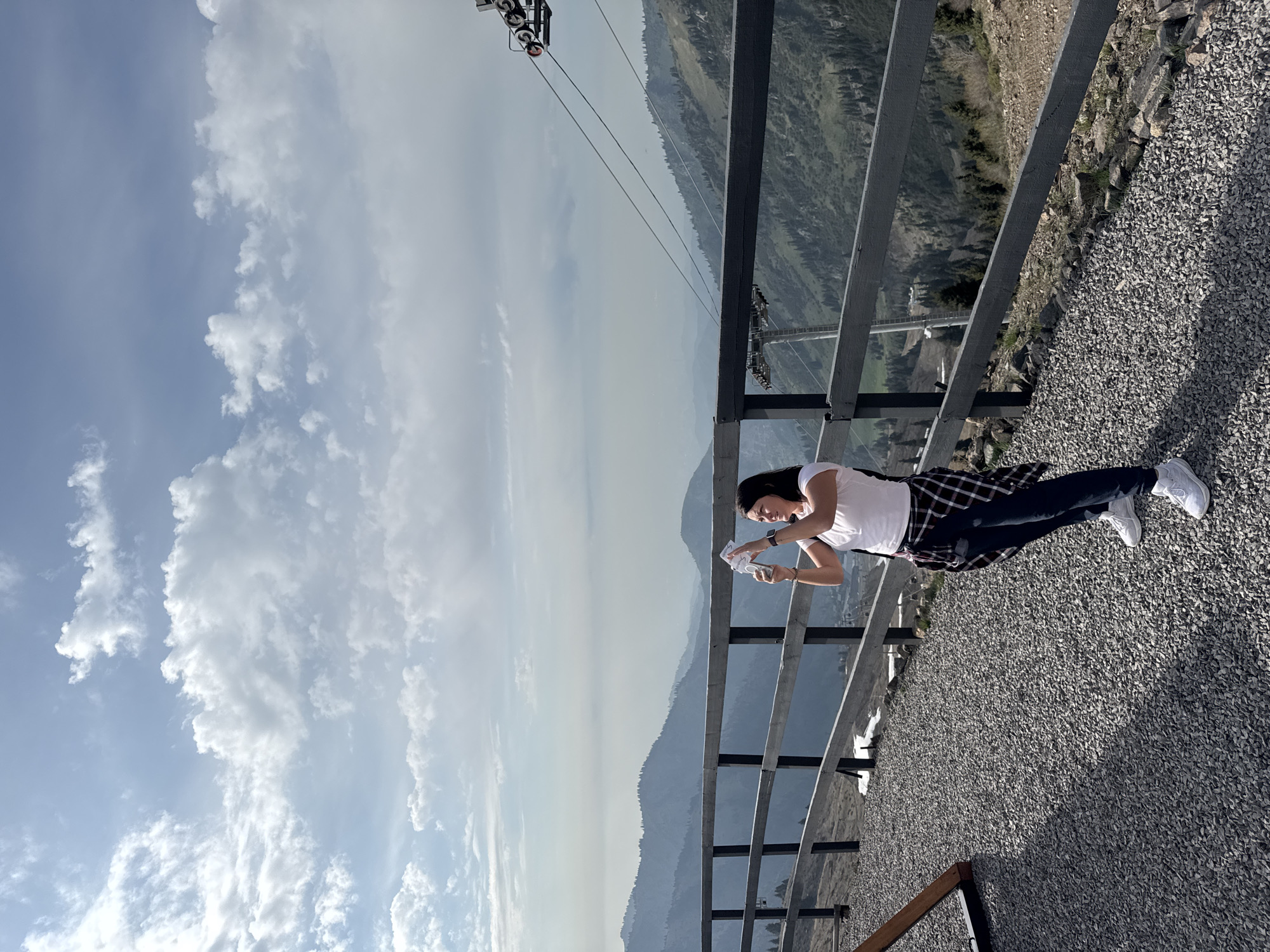 A woman is standing on a gravel surface near a metal railing, appearing to be examining or manipulating small white objects in her hands. She is wearing a white t-shirt, a dark plaid shirt tied around her waist, dark pants, and white sneakers. She has dark hair.
Behind the railing is a mountain landscape with visible hazy skies. A ski lift structure is visible in the upper portion of the frame. The location appears to be in a mountainous area.
The image is taken with a slight perspective, tilted. Given the location as Almaty, Kazakhstan, the scene could be on a mountain area in the Tian Shan mountains.