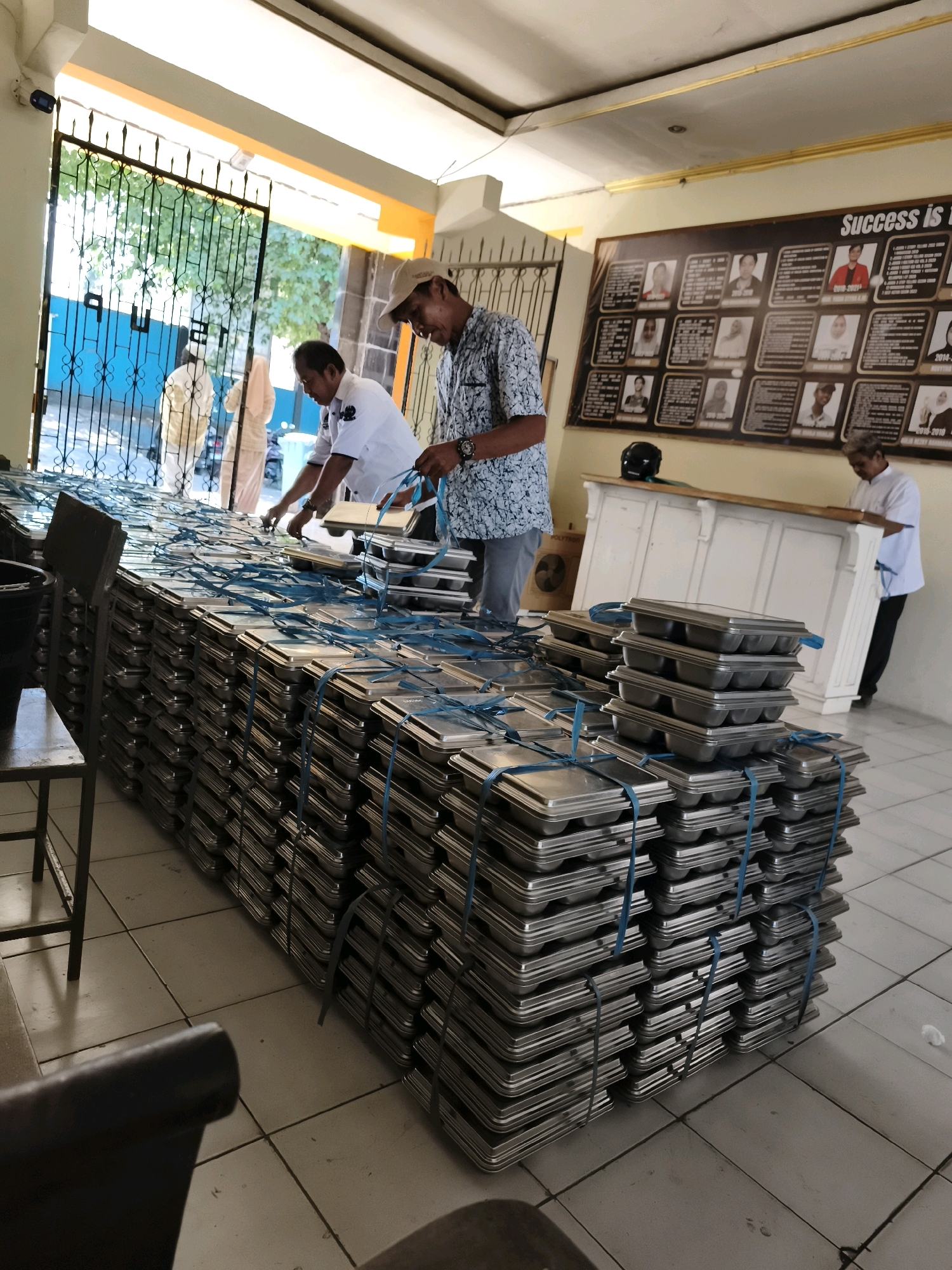 The image depicts a scene inside a building, possibly an office or community center, in Makassar, Indonesia. In the foreground, a large stack of metal trays, each secured with blue straps, dominates the view. Two men are visible interacting with these trays. One man, wearing a light-colored patterned shirt and a baseball cap, appears to be working with the trays, possibly organizing or preparing them. Another man, in a white uniform shirt, is also near the trays, his back to the camera. In the background, through an open gate, trees and foliage are visible, suggesting an outdoor area beyond. A bulletin board with photos and text is mounted on the wall, with the word "Success is" visible, indicating it might be an achievement board or recognition display. The overall environment appears to be a functional space, possibly a distribution point for the trays.